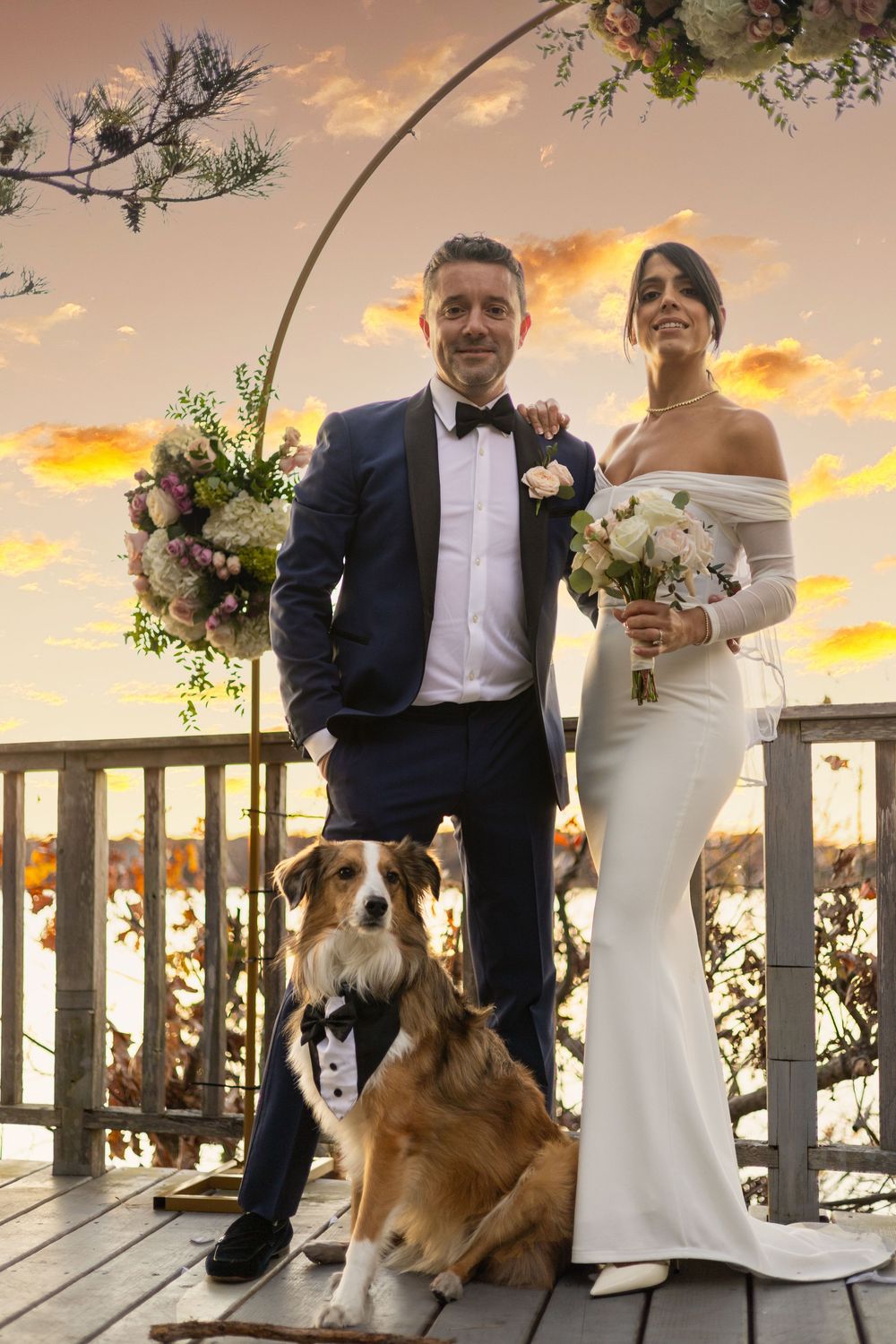 A couple poses with their dog at a sunset wedding ceremony on a white porch decorated with floral arrangements.