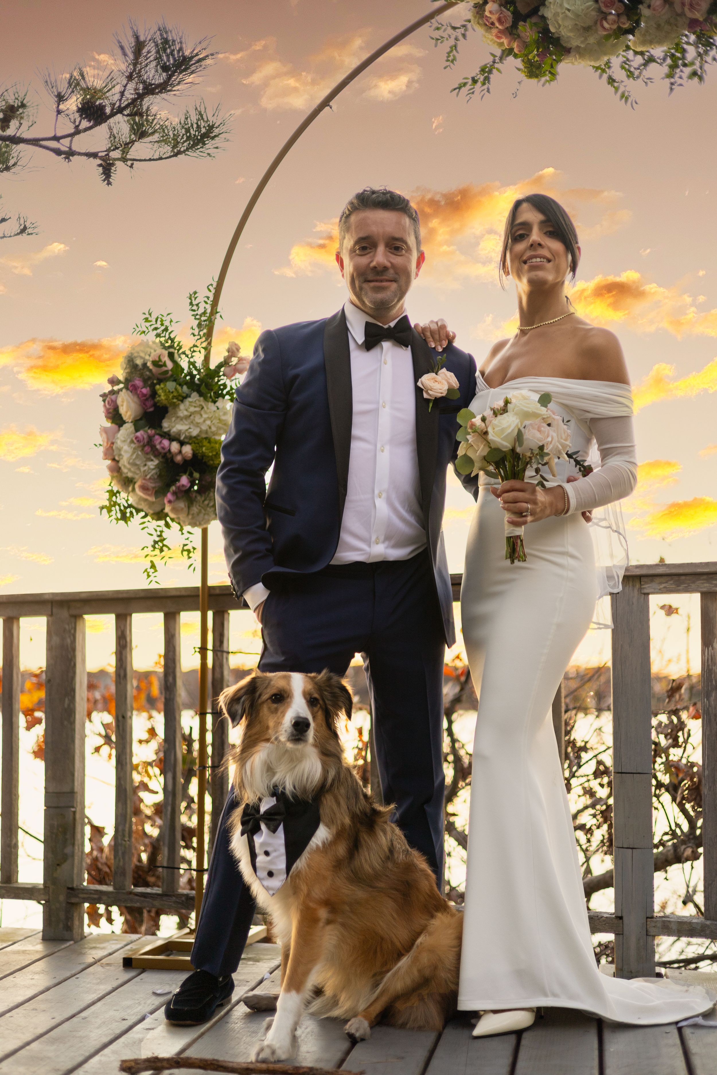 A couple poses with their dog at a sunset wedding ceremony on a white porch decorated with floral arrangements.