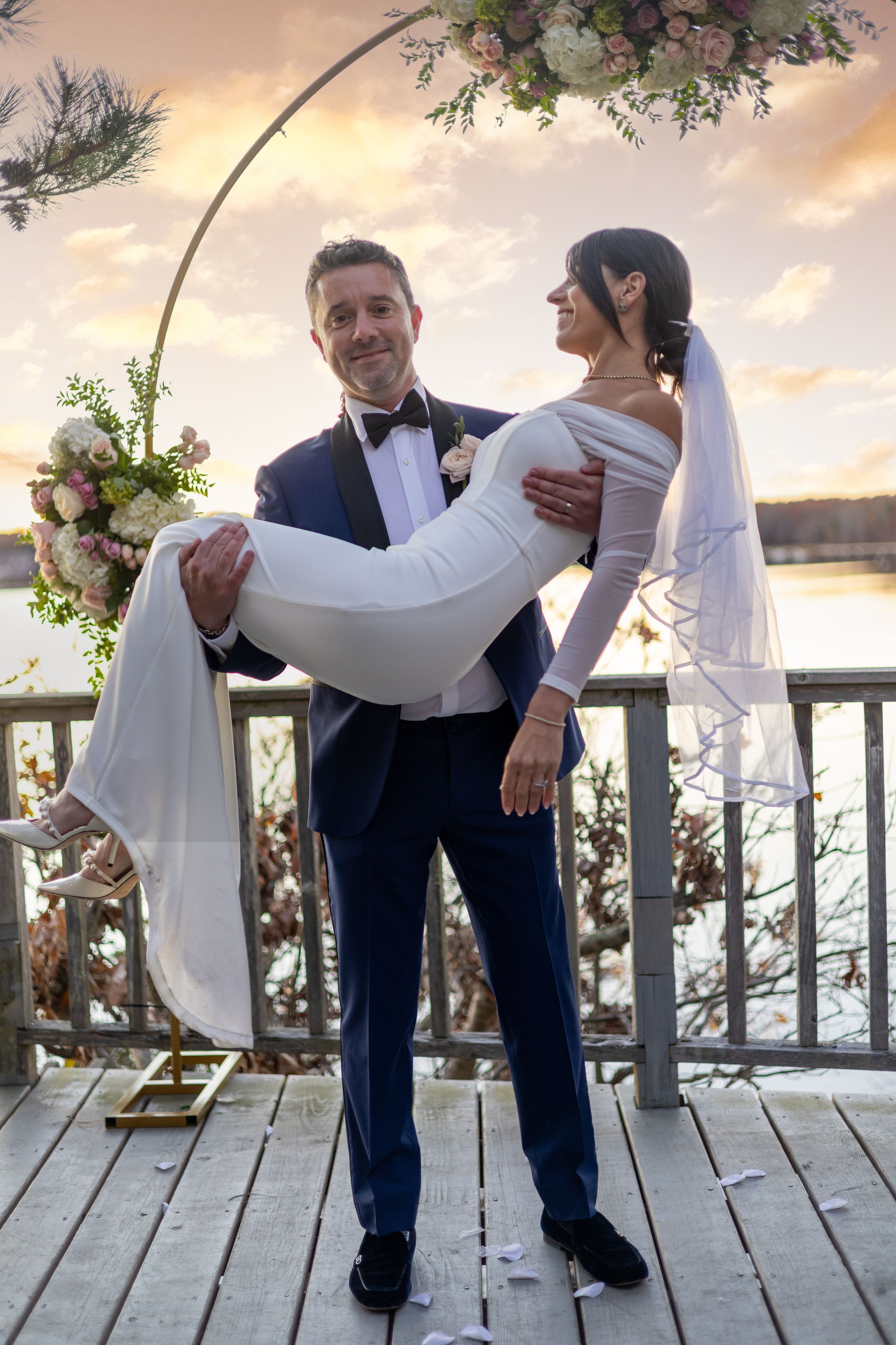 A romantic wedding couple poses on a wooden deck at sunset, with floral arch decorations and a waterfront backdrop.