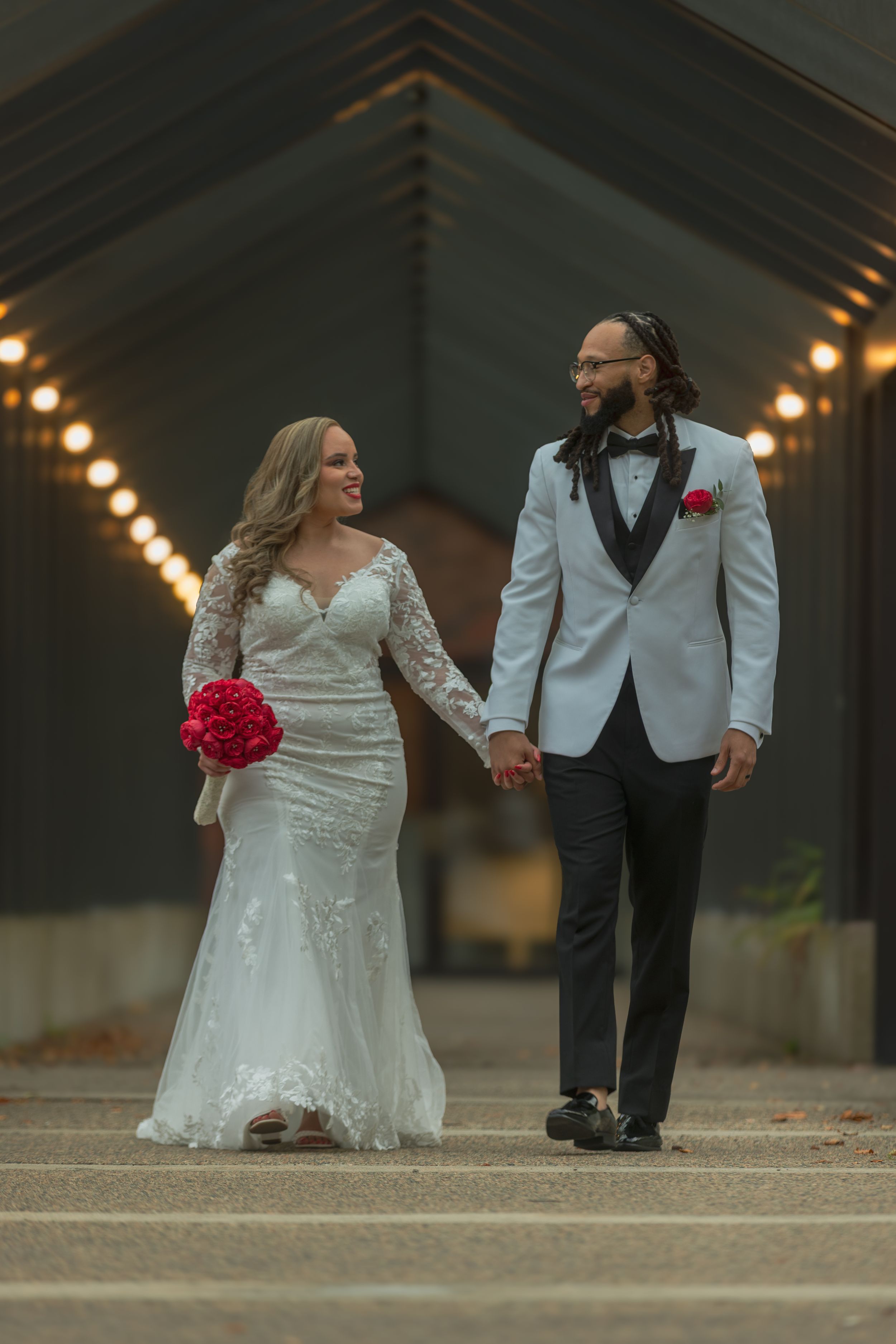 A bride and groom share a playful moment walking together outdoors with string lights and red bouquet details.