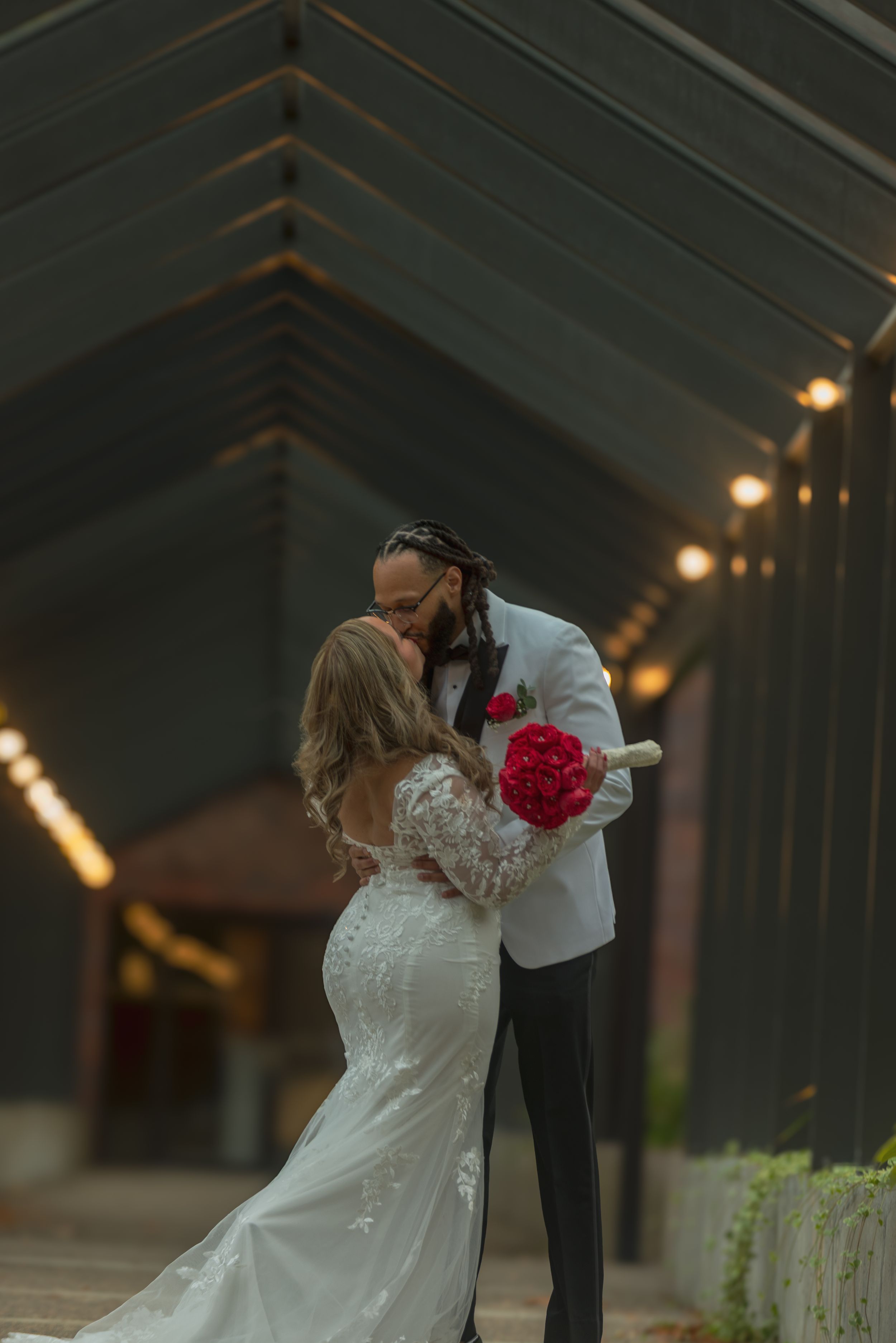 A couple shares a romantic kiss under a rustic wooden pavilion decorated with soft lighting.