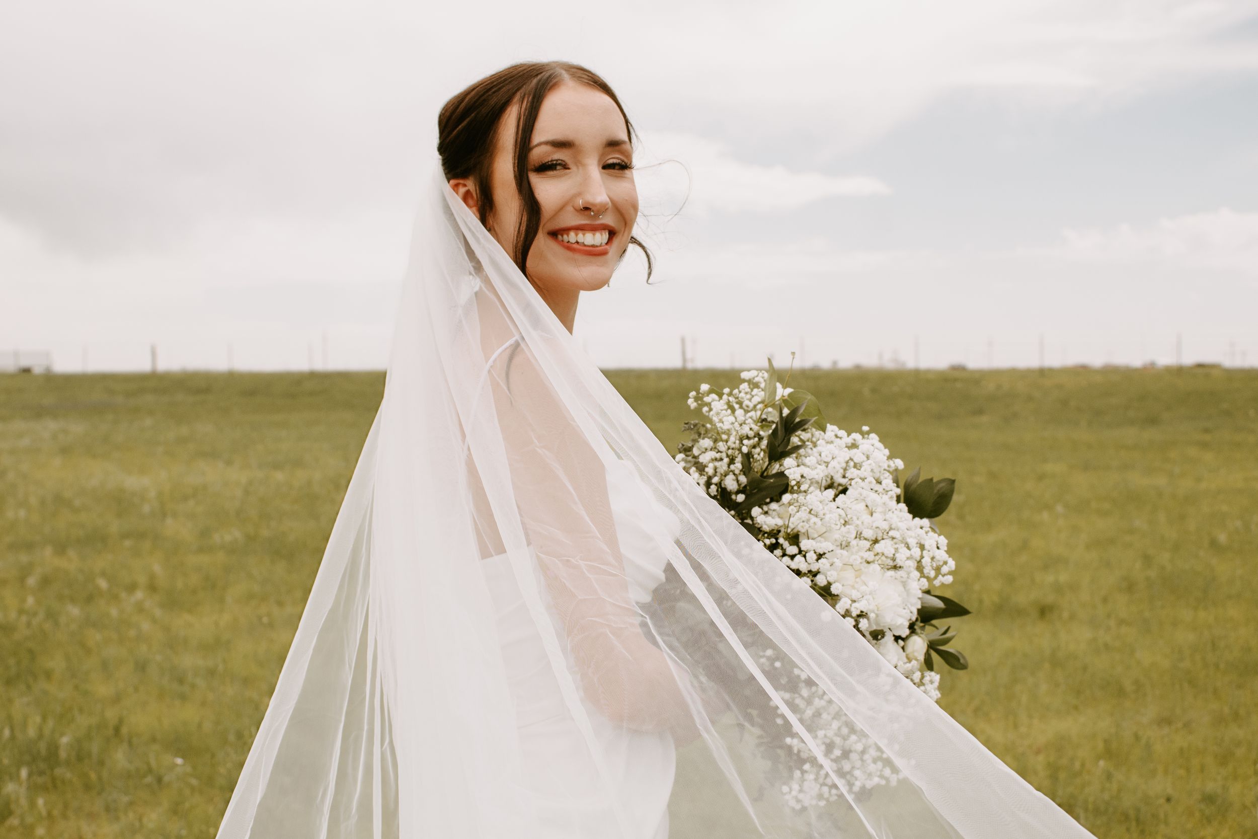 Smiling bride in white gown and veil holding bouquet of white flowers in a green field.
