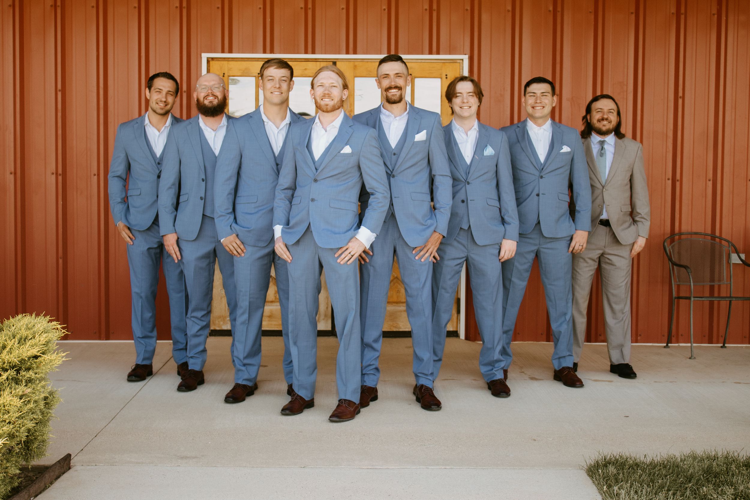 Groomsmen in matching light blue suits pose with groom outside a rustic barn wedding venue.