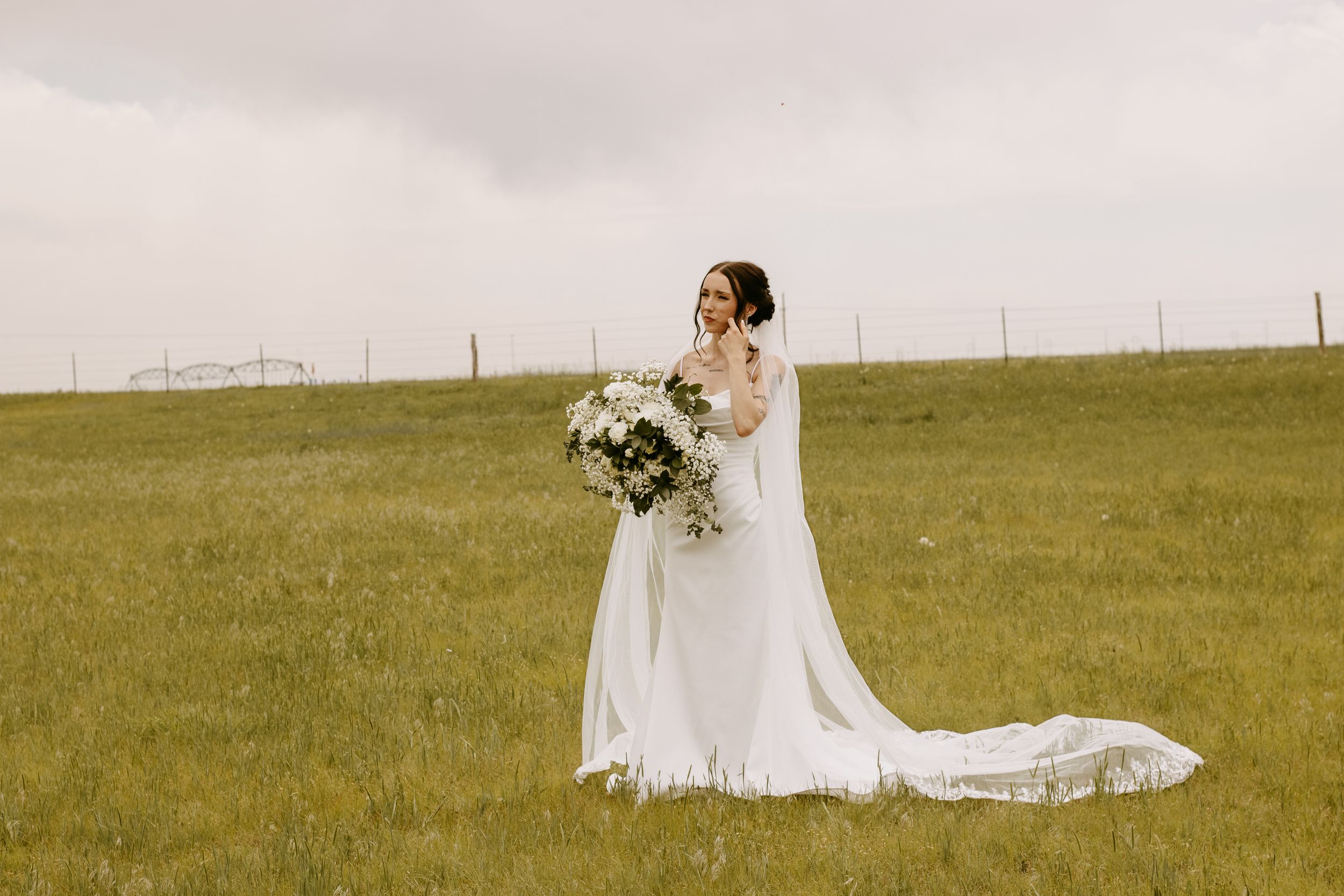 Bride in white gown with long train holds bouquet in green field under cloudy sky.