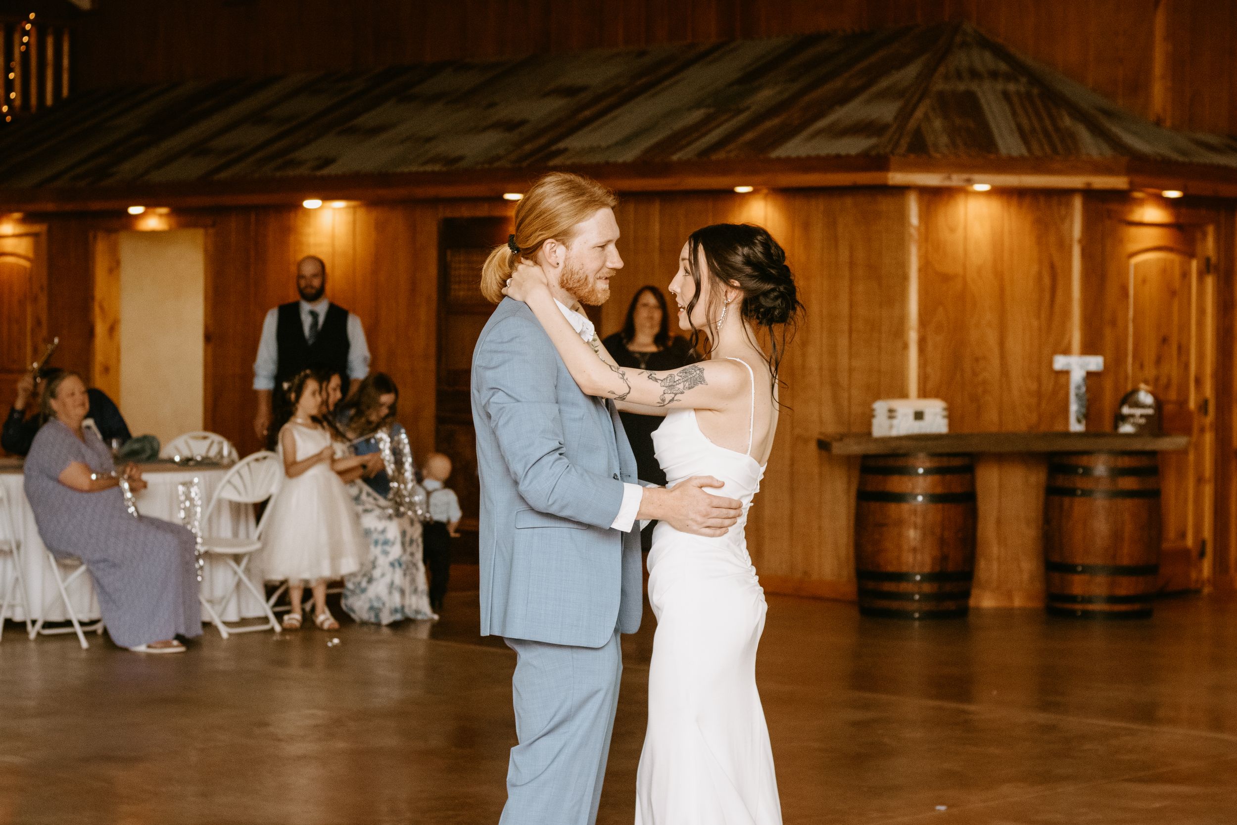 Bride and groom share their first dance in a rustic wood-paneled reception hall with guests seated at tables.