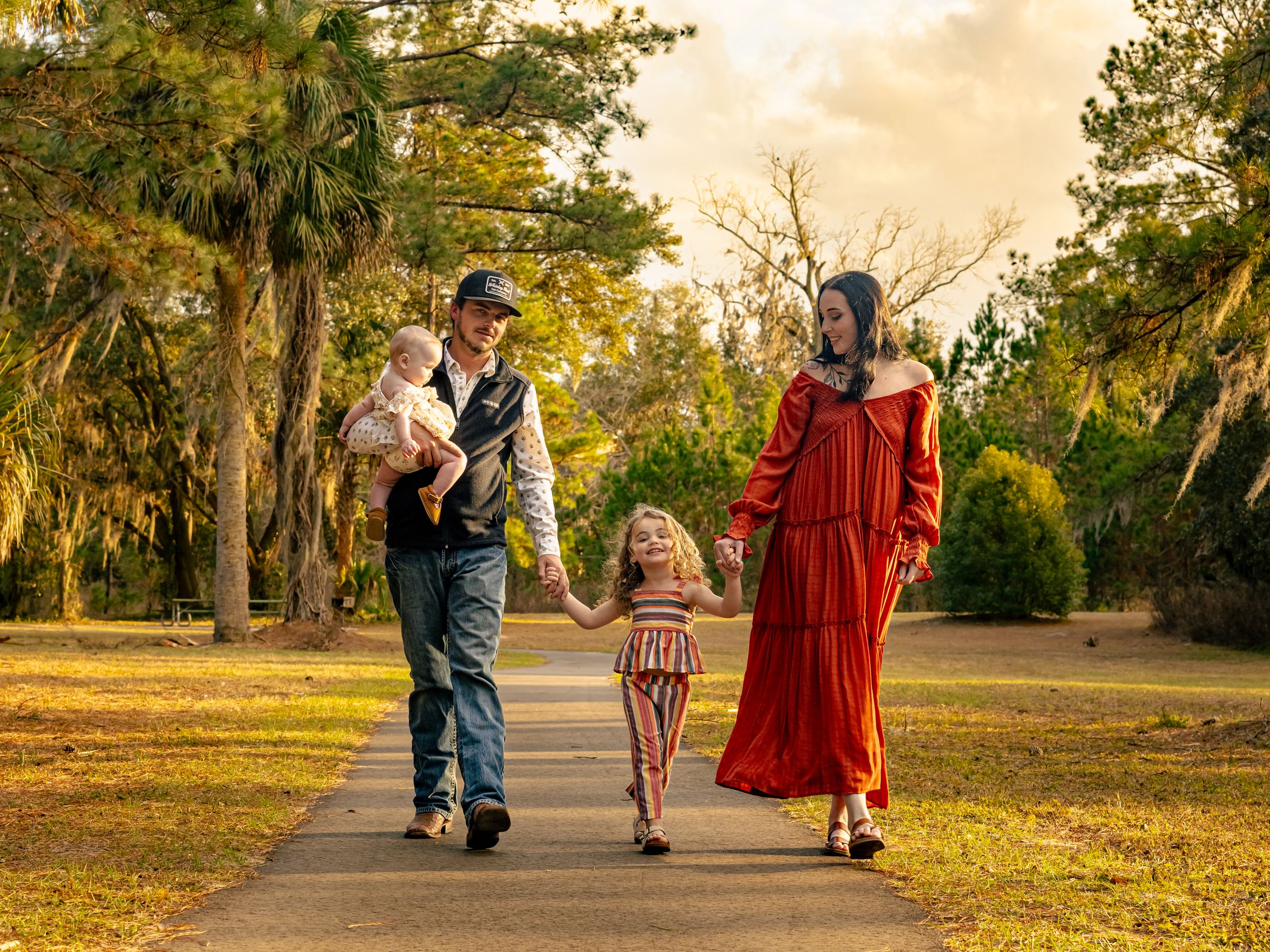Family of four walks hand-in-hand along a sunlit park path at sunset.
