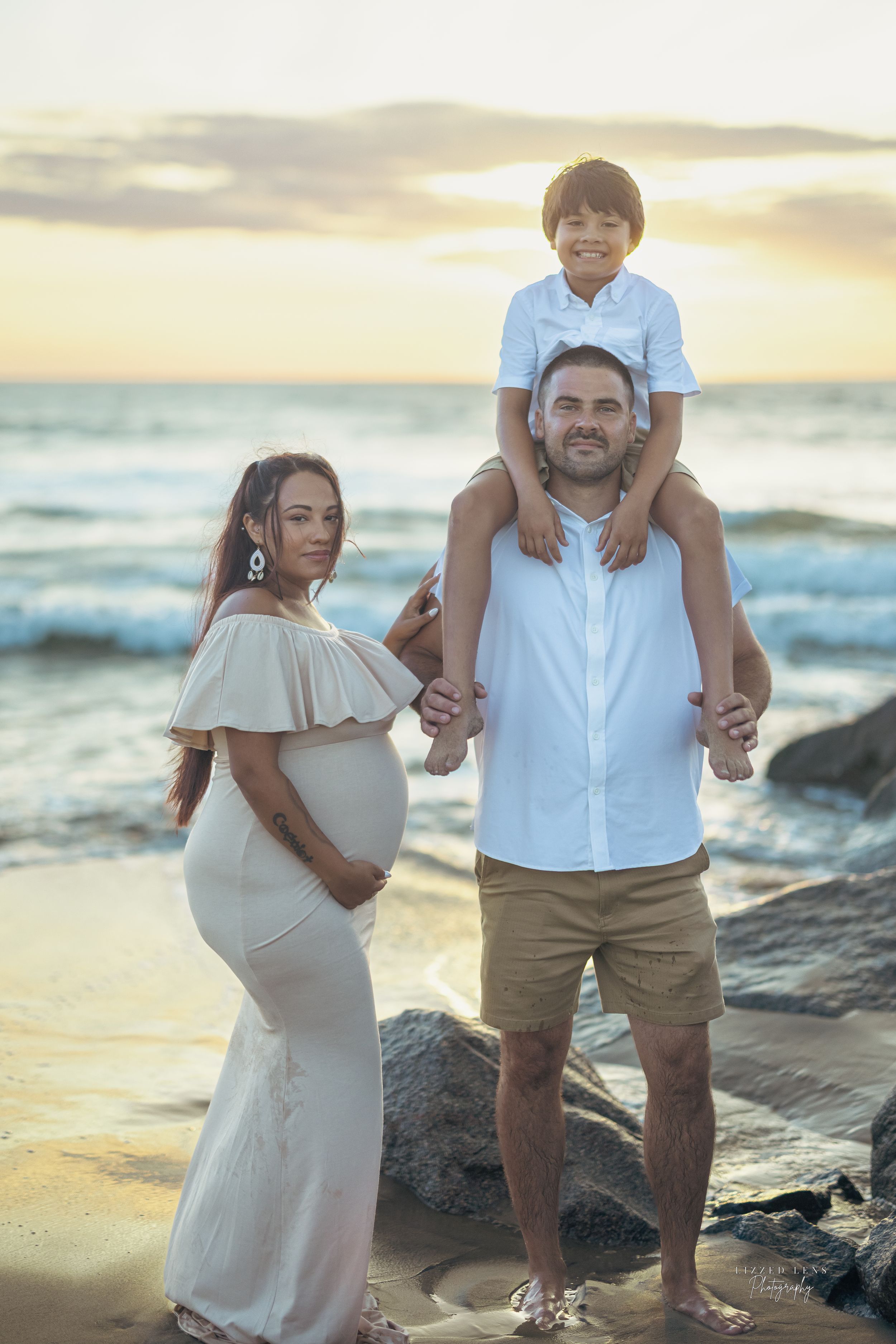 Pregnant woman in white dress with husband and son at beach sunset.