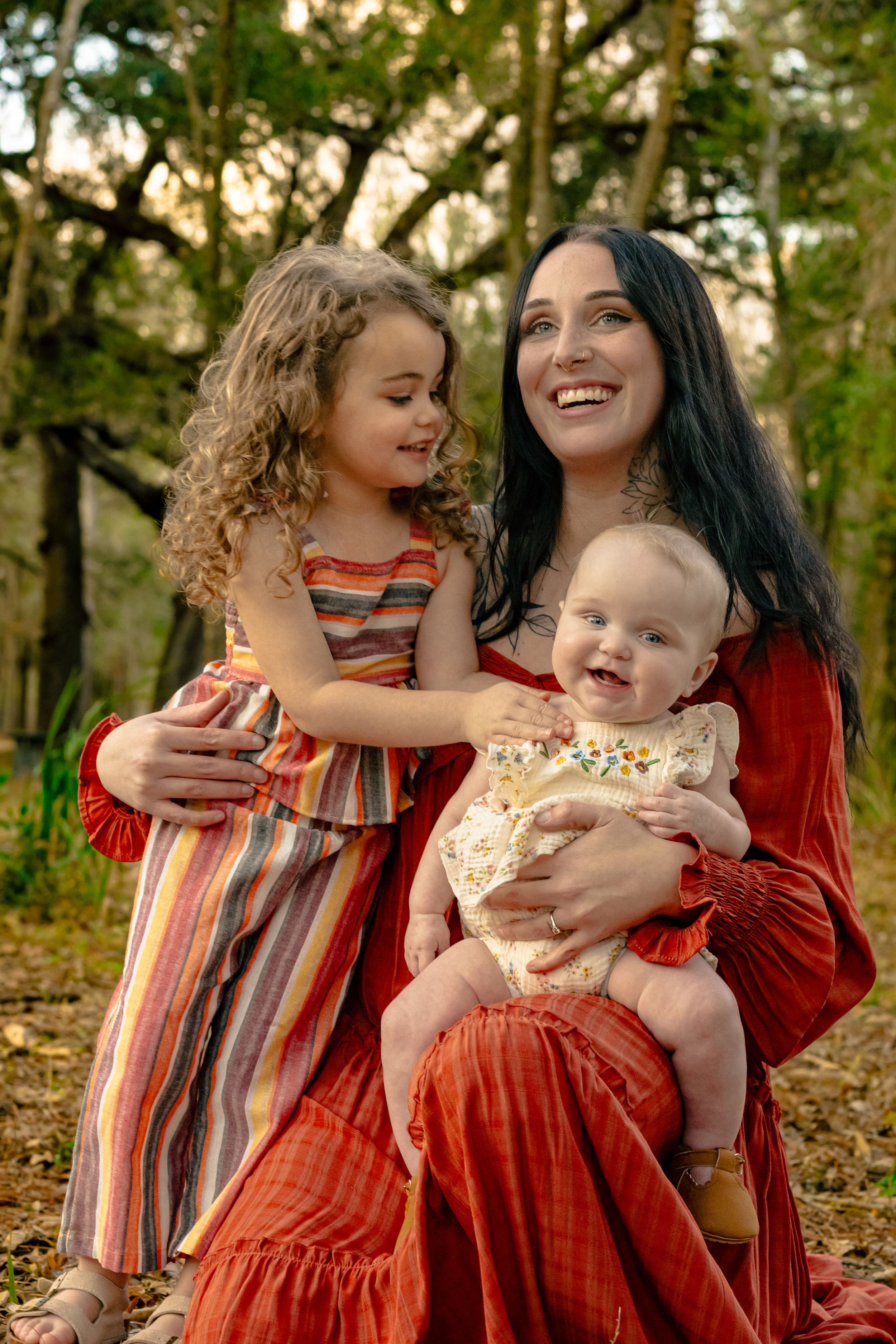 Smiling mother in red dress sits with baby and curly-haired daughter in park.