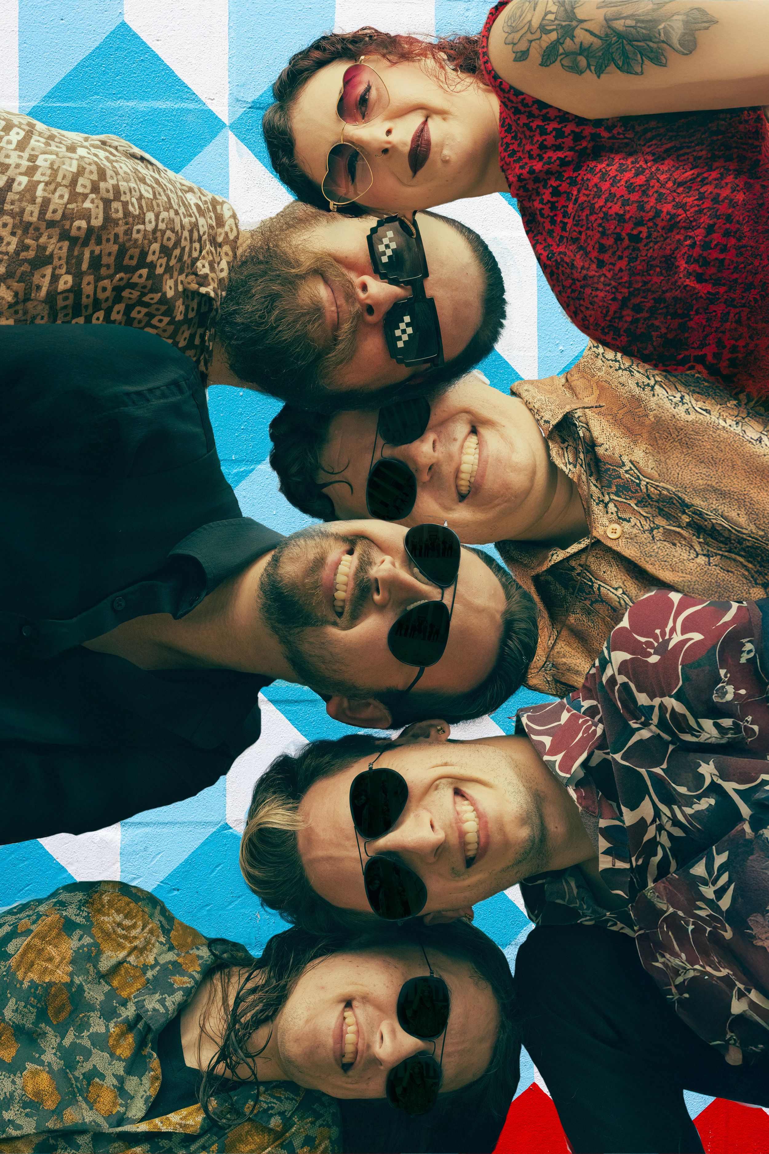Seven smiling band members huddle together wearing sunglasses against a blue geometric wall.