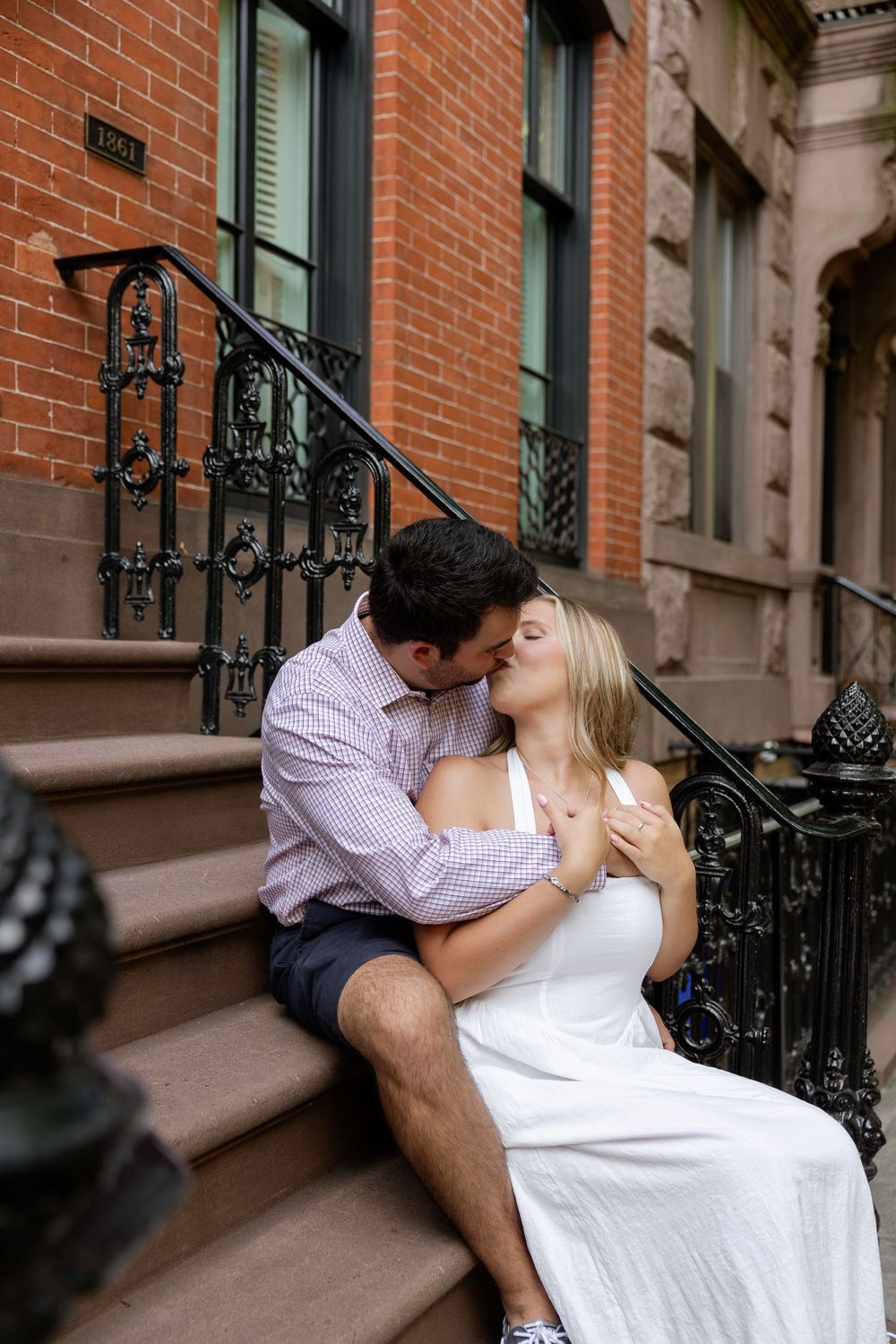 A romantic couple shares tender kisses while sitting on brownstone steps in an urban neighborhood.