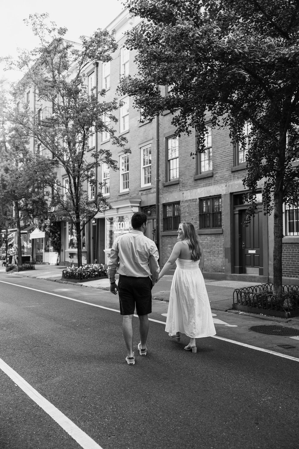 Two people walking down a quiet tree-lined residential street with brick townhouses and parked cars.
