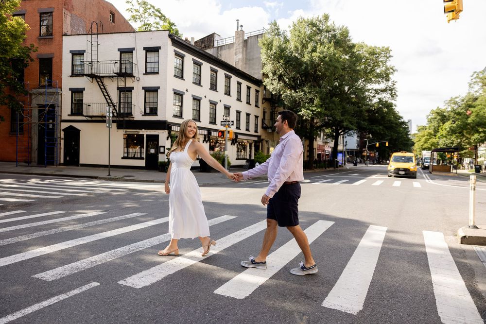 A couple walks across a city crosswalk in white summer outfits during a sunny day in an urban setting.