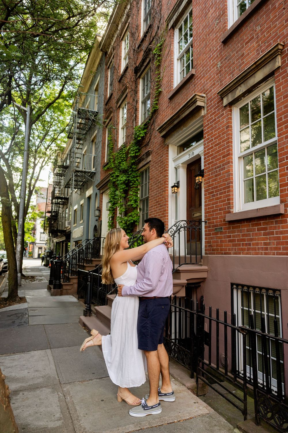 Couple sits on brownstone steps in a historic neighborhood with brick buildings and iron railings on a sunny day.