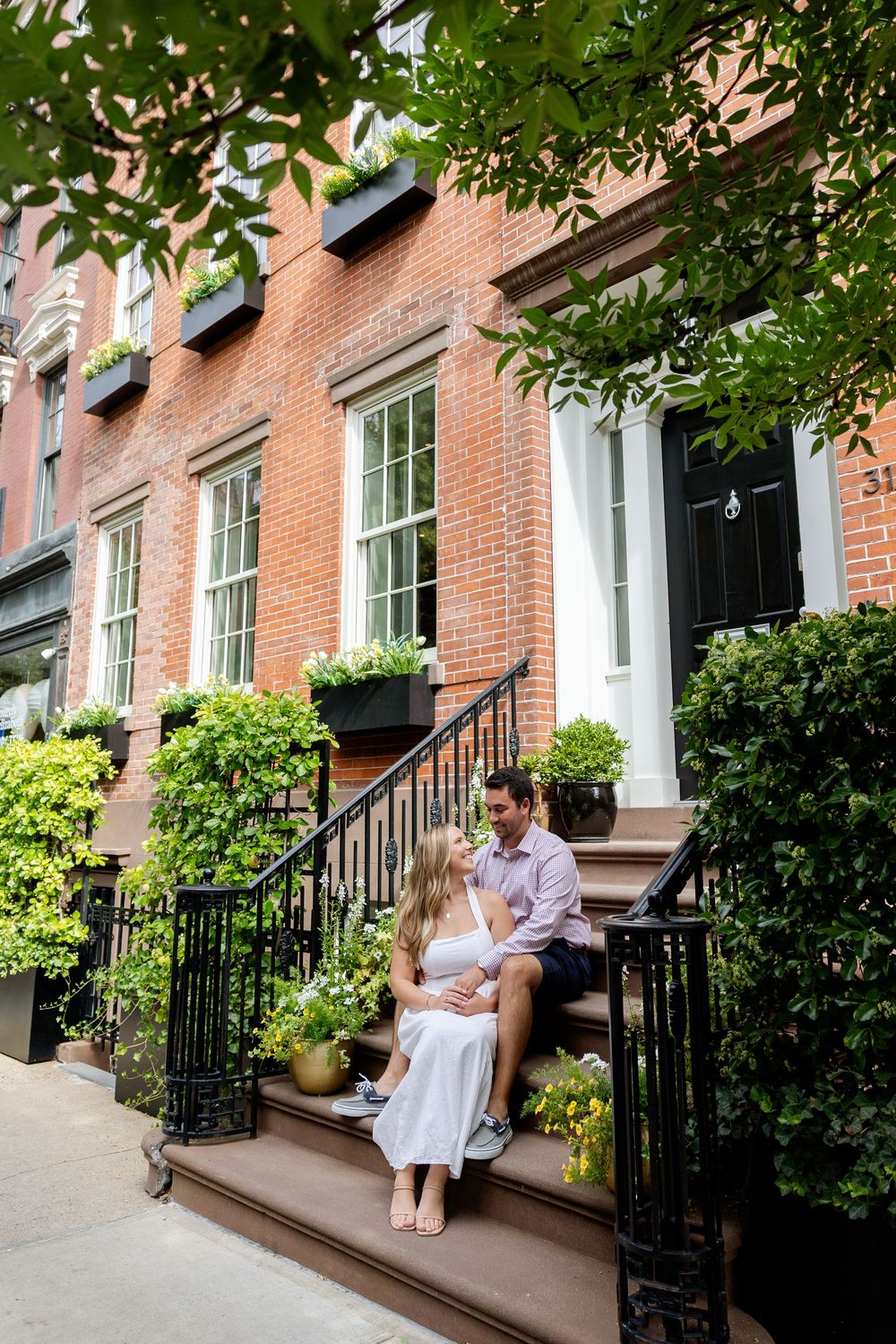 Two people relaxing on front steps of brick townhouse with black railings and lush greenery overhead.