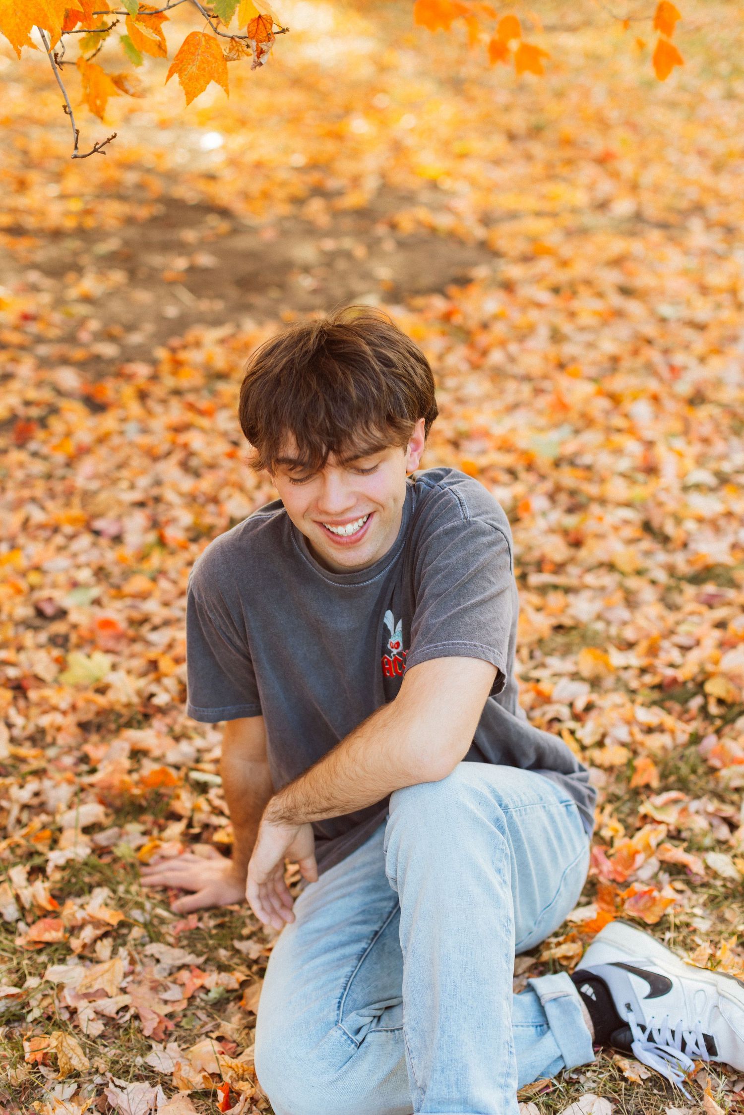 A young man sitting on a bed of fallen golden and orange leaves laughs and looks down during a Kansas City senior photography session.