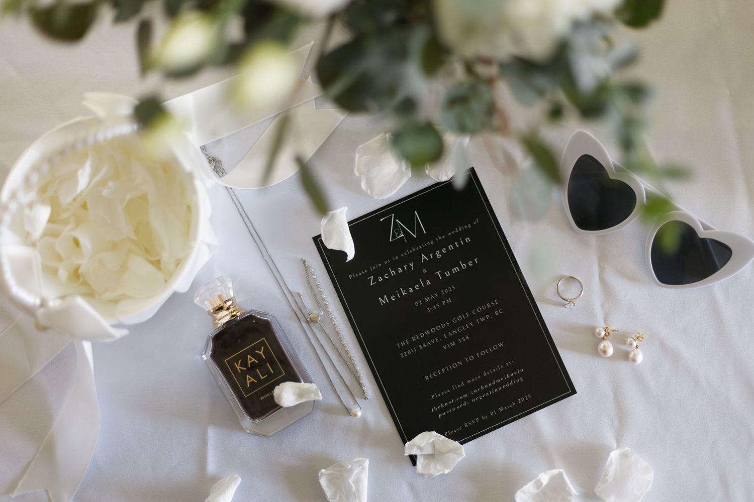 Elegant black and white wedding invitation suite with florals, perfume, sunglasses, and pearl accessories flatlay.