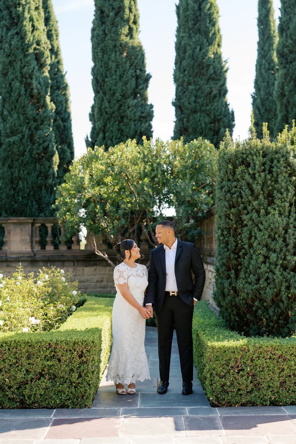 A couple walks and poses together in a garden with tall cypress trees and manicured hedges on a sunny day.