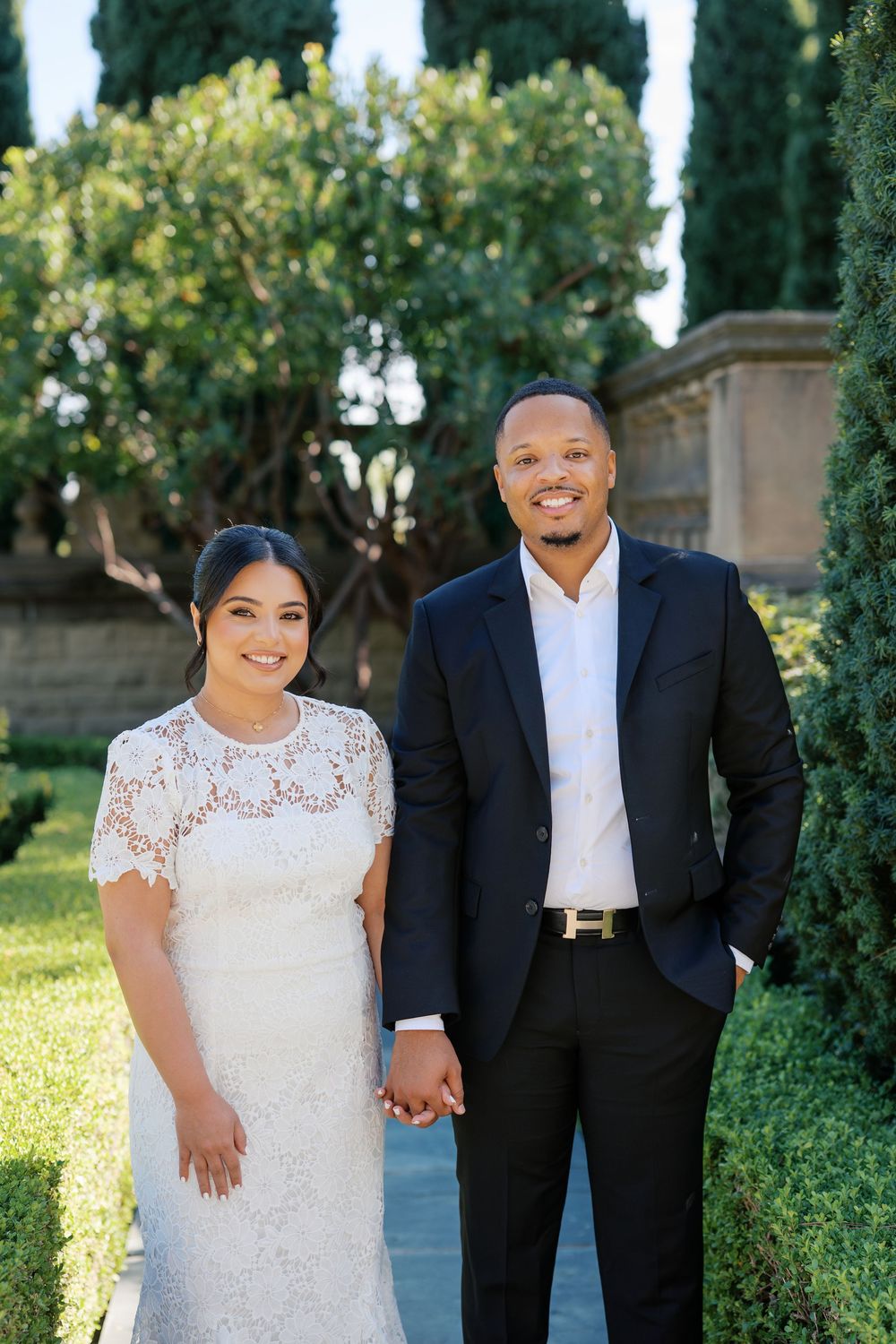Wedding couple poses together on stone path with cypress trees creating elegant garden backdrop.