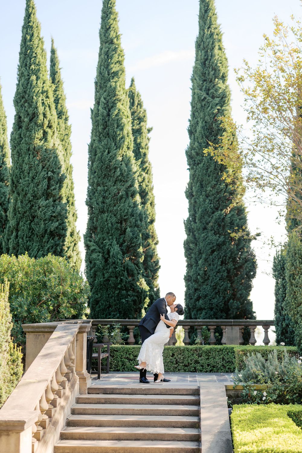 Elegant couple embraces on grand stone staircase surrounded by tall cypress trees in formal garden setting.