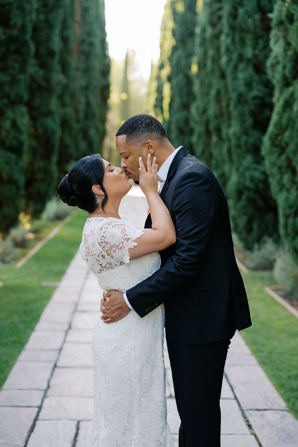 A romantic wedding couple shares intimate kisses along a cypress tree-lined path in an outdoor garden setting.