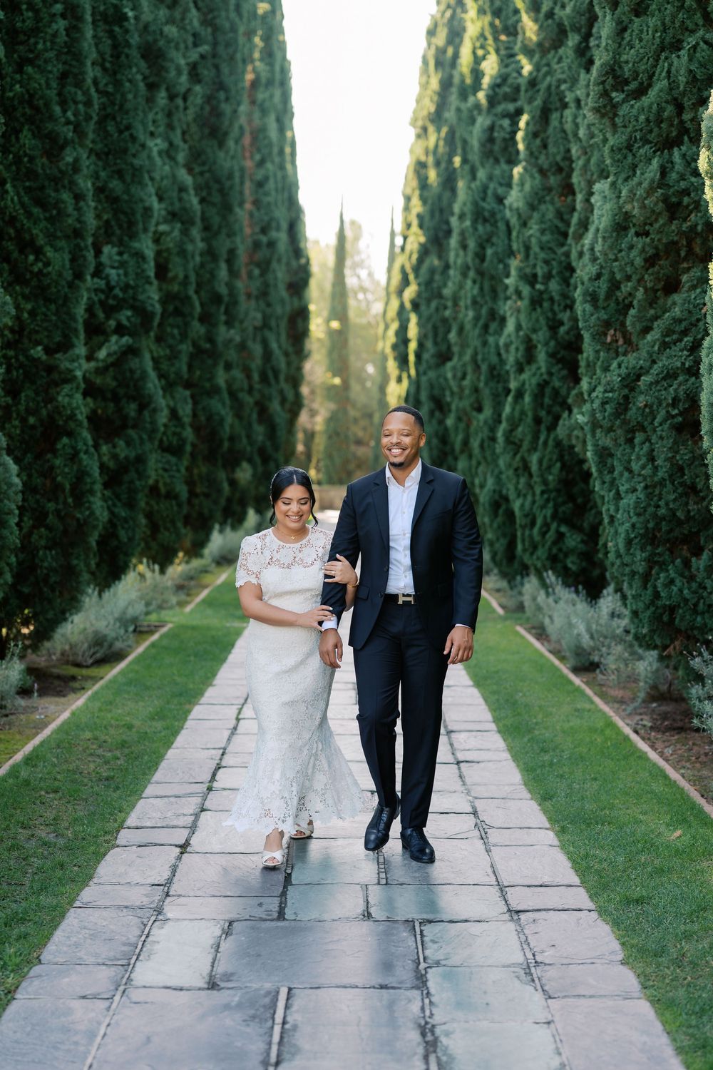 Newlyweds share playful moments while walking along stone pathway lined with tall cypress trees.