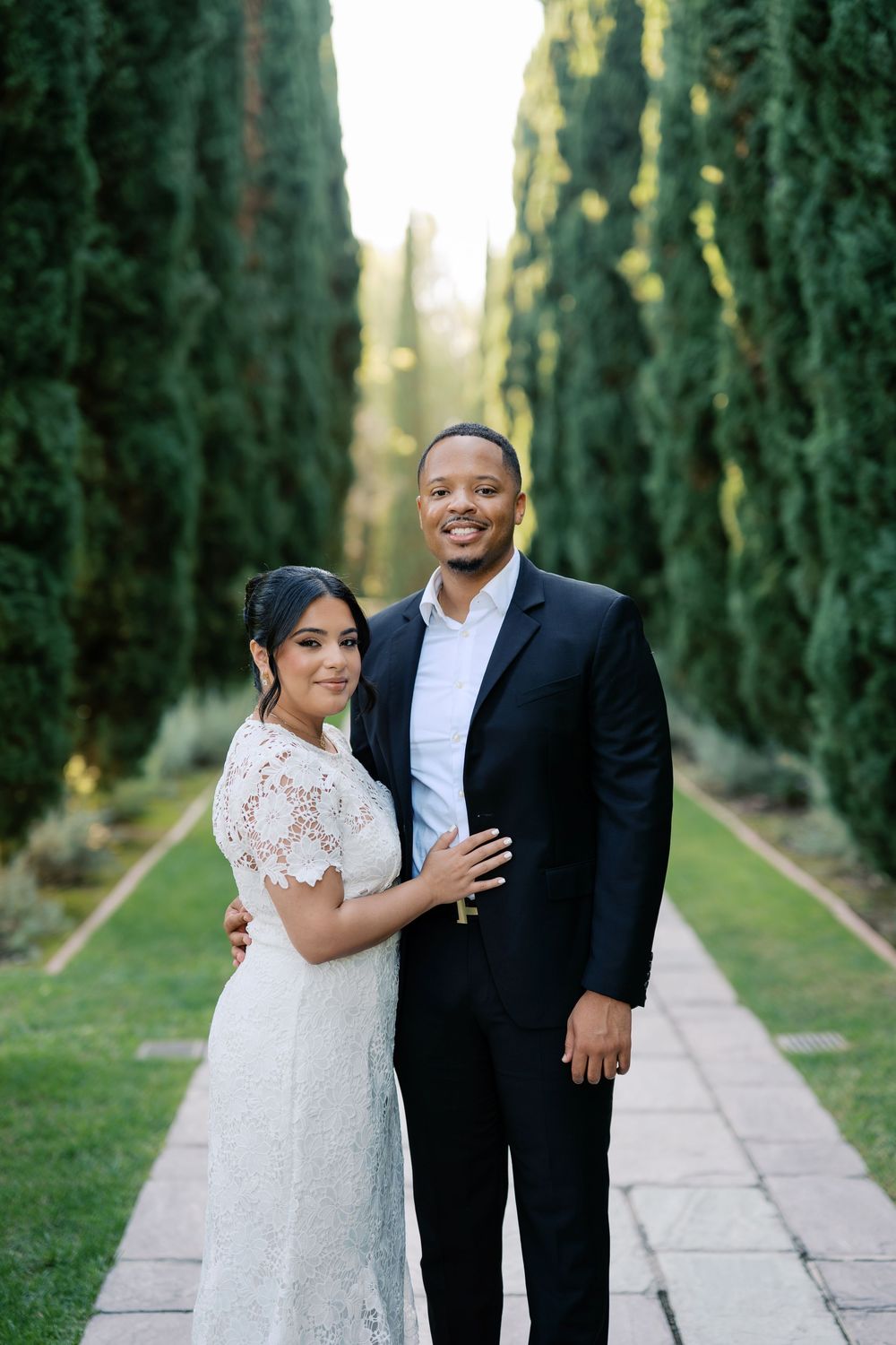 Happy couple embraces on garden path surrounded by majestic cypress trees in formal attire.