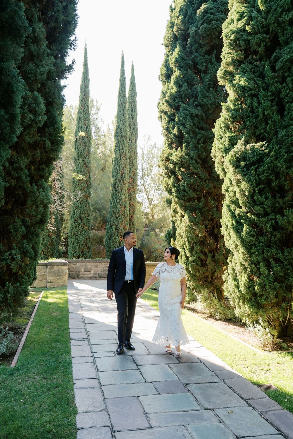 A couple walks along a scenic path lined with tall cypress trees in a Mediterranean garden setting.