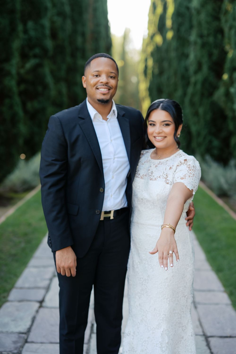 A couple poses together in a garden path lined with tall cypress trees, the bride wearing a white lace gown.