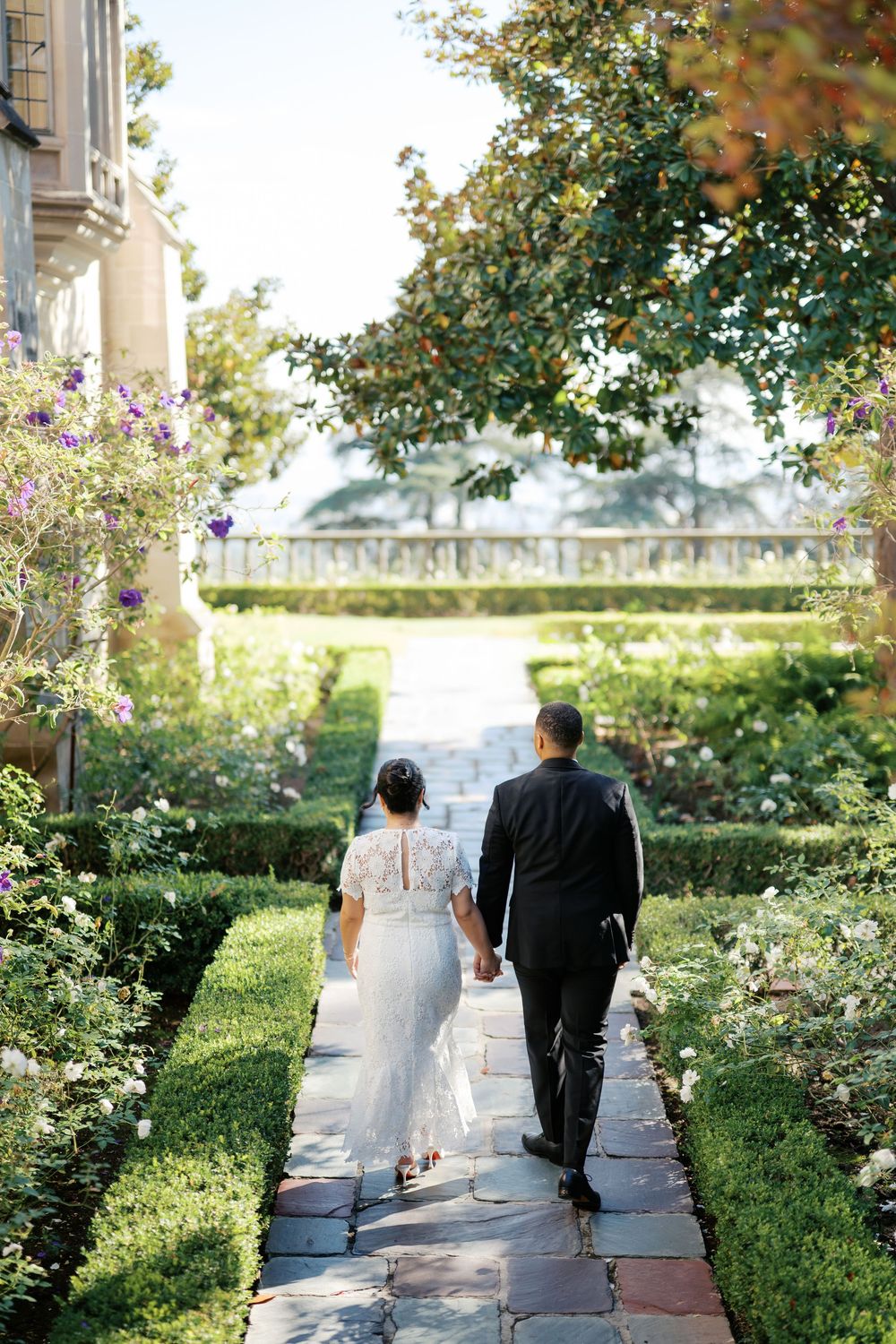 A couple walks along a garden path surrounded by lush greenery and stone architecture on a sunny day.