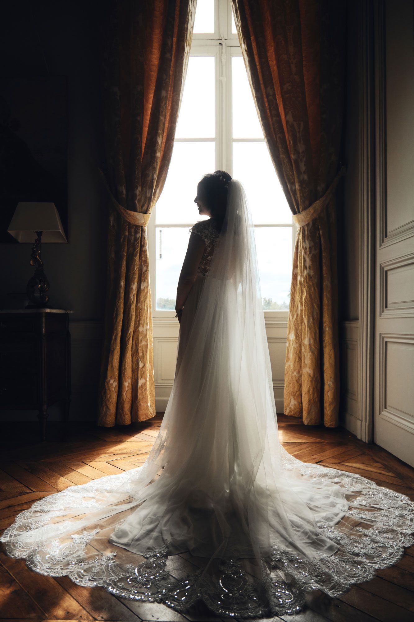 A silhouette of a bride in a flowing wedding gown stands by a window with elegant curtains.