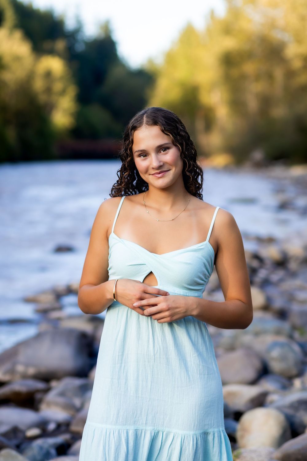 A person in a light blue sundress poses outdoors near a rocky riverbank on a sunny autumn day.