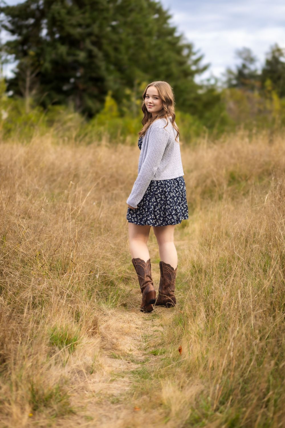 Person in brown boots and floral skirt walking through a golden field of tall grass on a sunny autumn day.