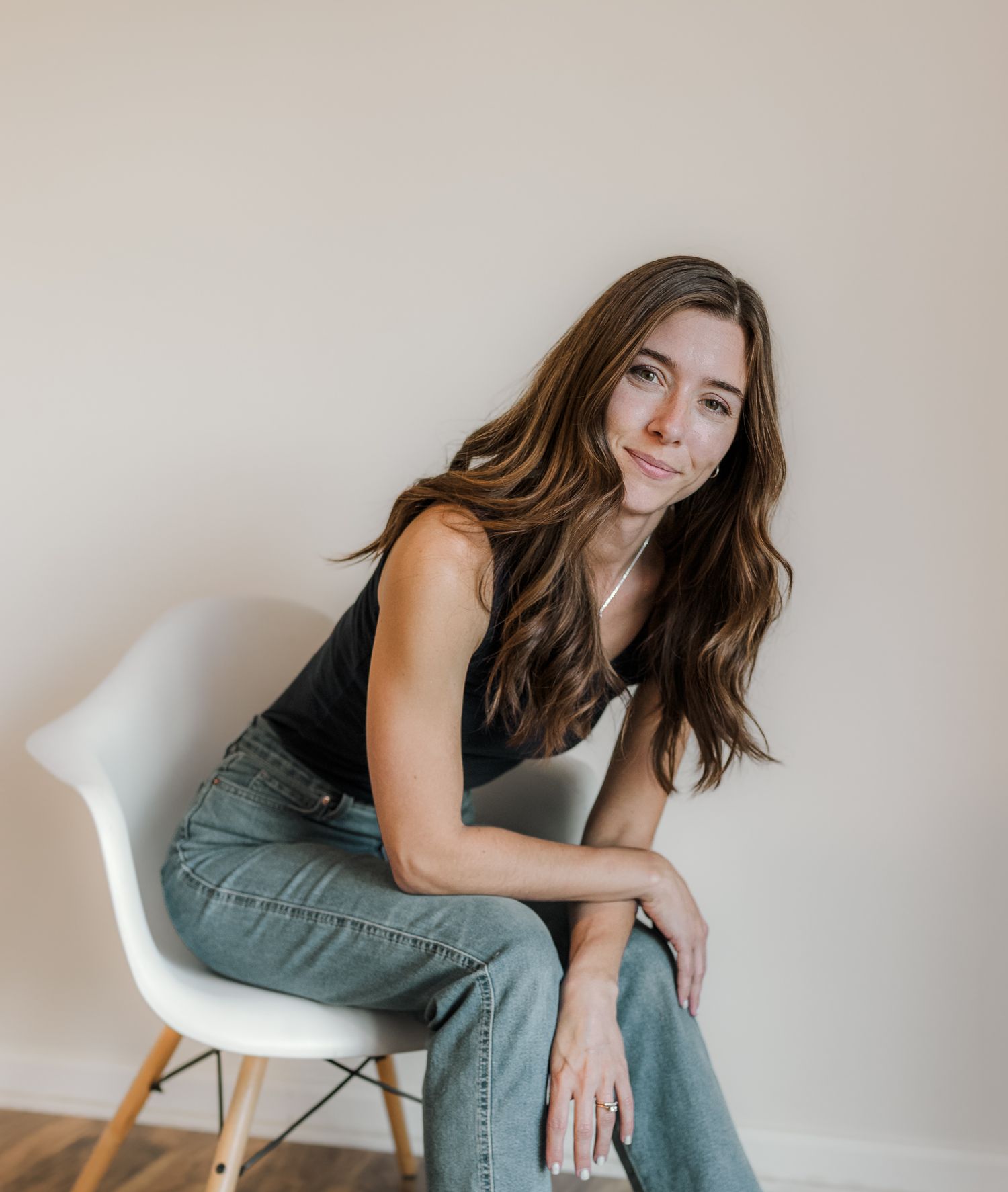 A person with long brown hair wearing a black tank top and jeans sits casually in a white modern chair.