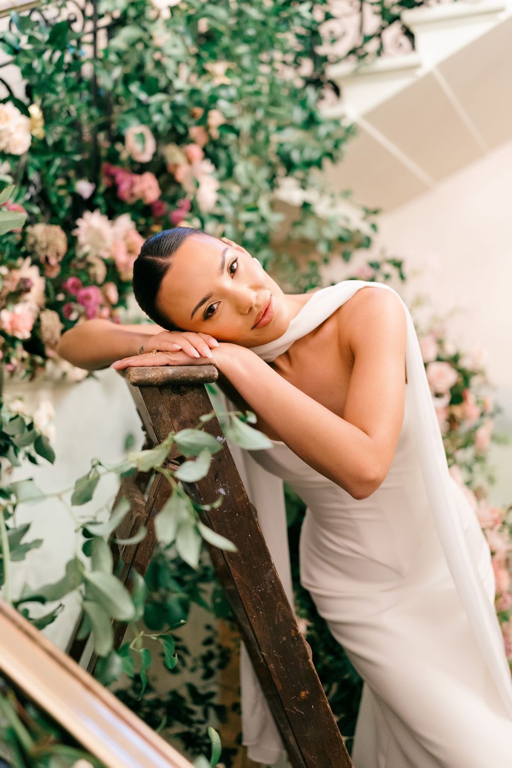 A person in a white sleeveless dress poses gracefully against a backdrop of lush greenery and pink flowers.