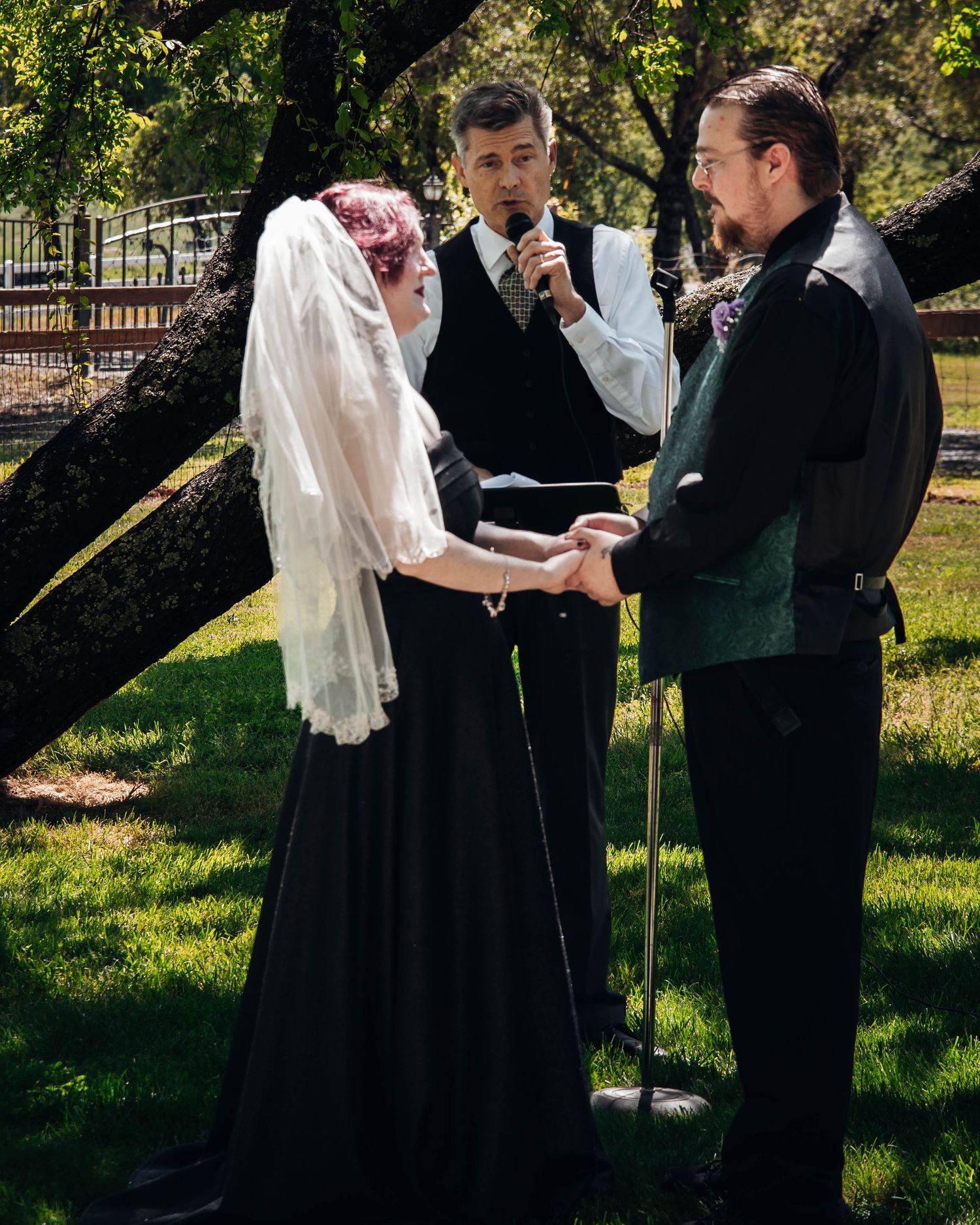 Wedding ceremony taking place outdoors under trees with guests watching the proceedings.