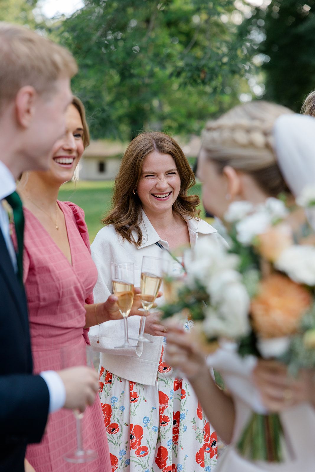 A group of wedding guests in pink and floral dresses laughing and socializing while holding drinks at an outdoor reception.