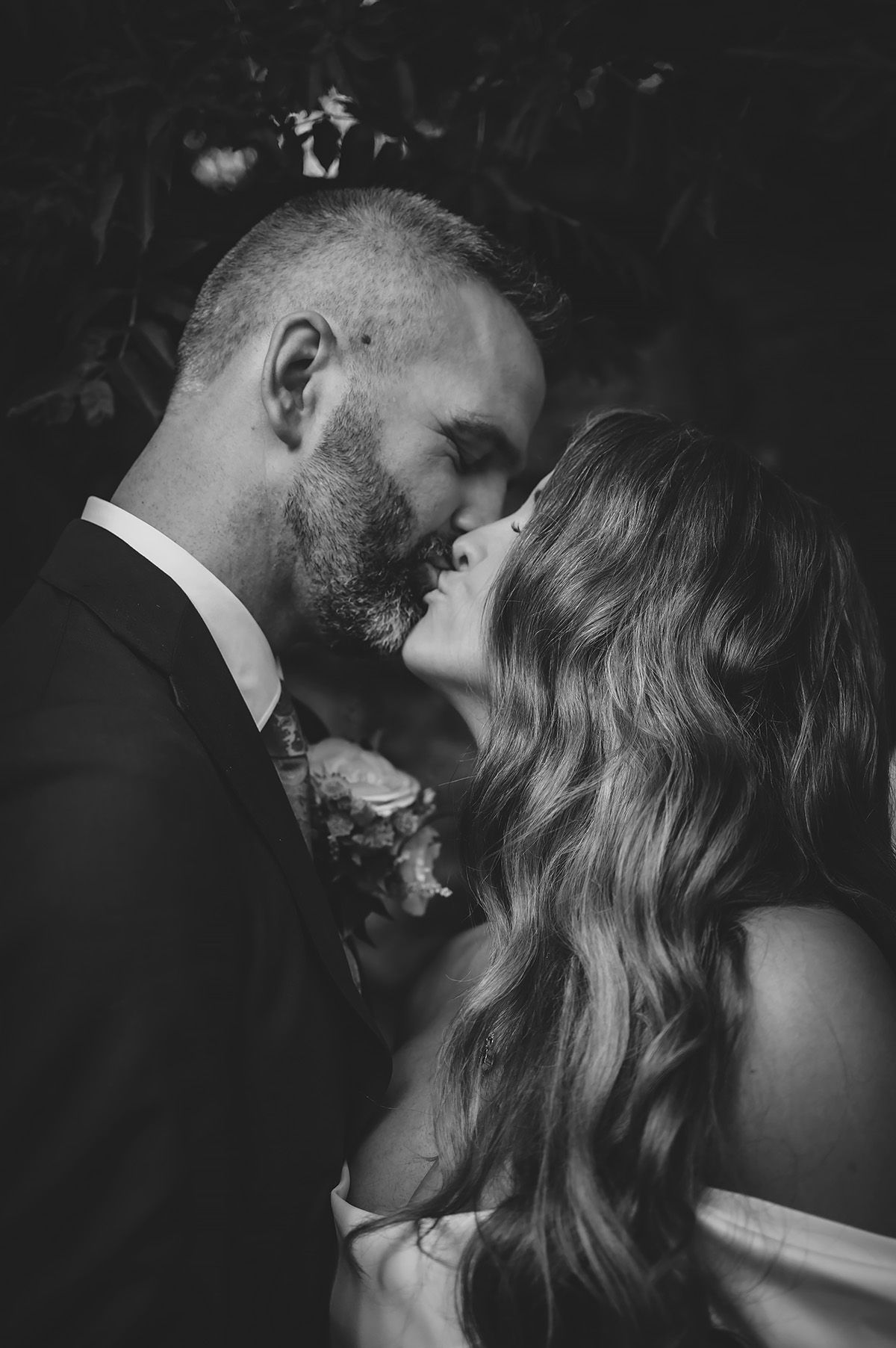 Nära porträtt i svartvit av ett brudpar som möts i en öm kyss. Bröllopsfoto med edge, mjuk konstrast och filmisk känsla. Fotograf Lets Bloom