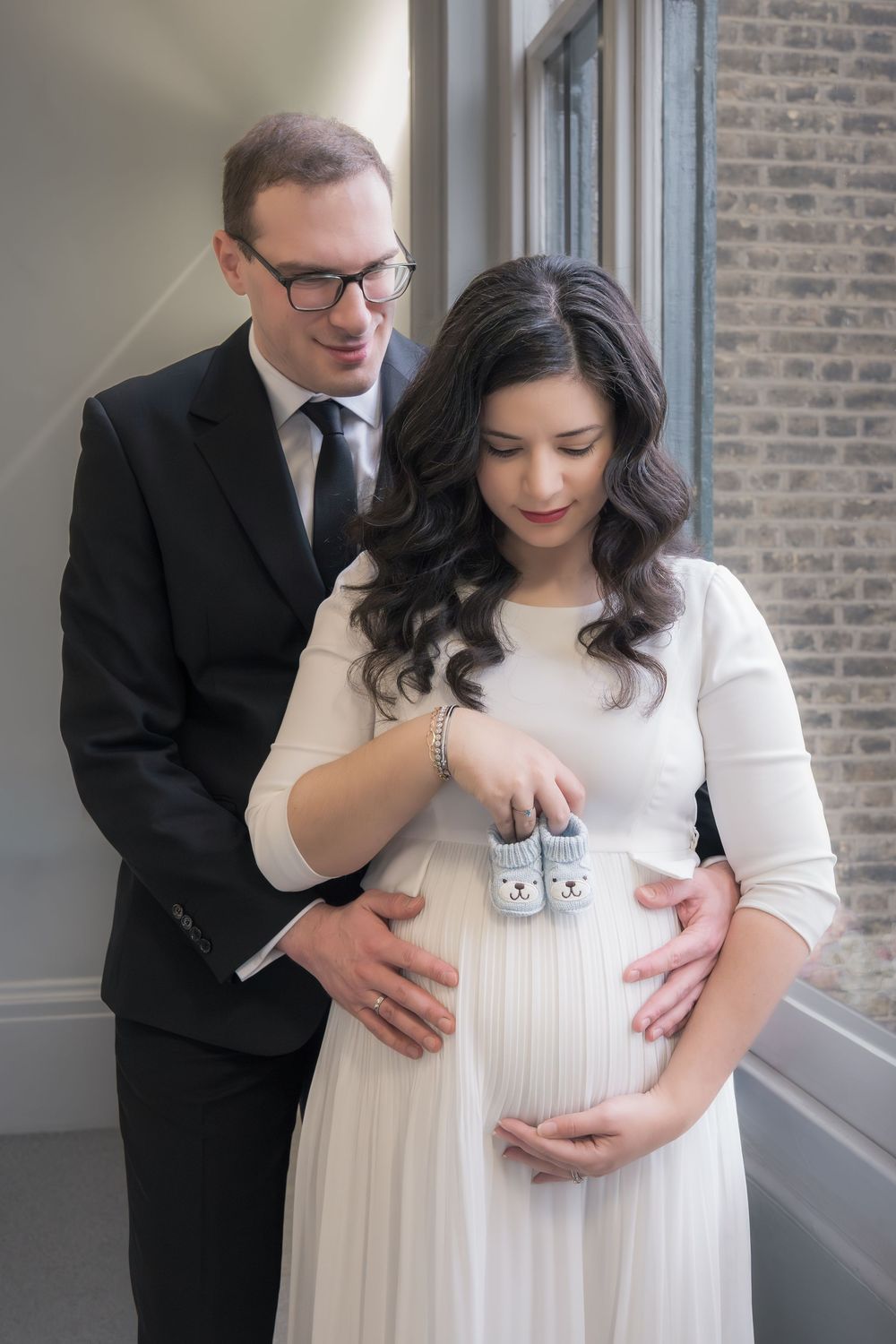 Expectant couple holds tiny baby shoes while posing near window in formal attire during maternity photoshoot.