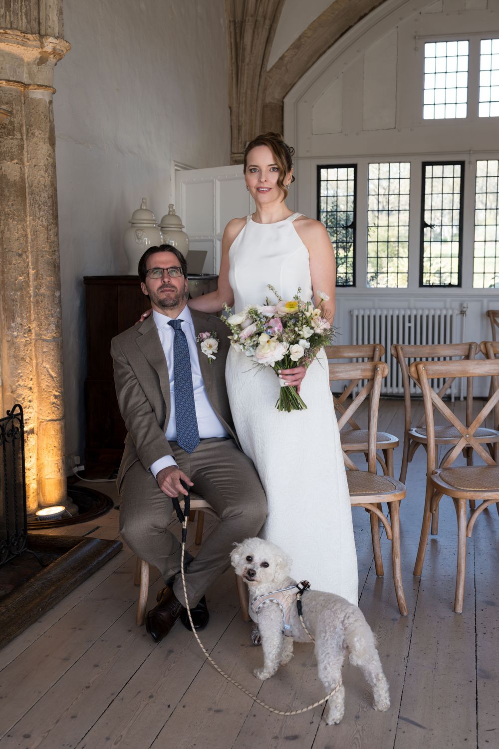 Bride and groom pose with white fluffy dog inside elegant wedding venue with wooden floors and leaded windows.
