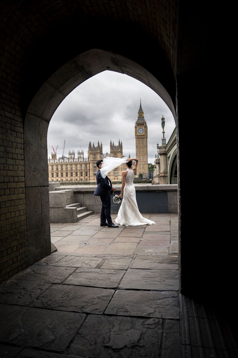 Bride and groom pose under stone arch with Big Ben and Houses of Parliament in London backdrop.
