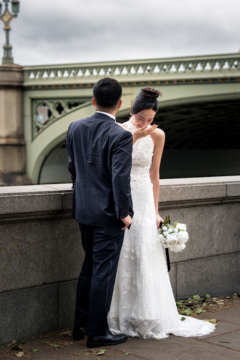 Bride and groom pose by a stone wall near Westminster Bridge in London, bride holding white bouquet.