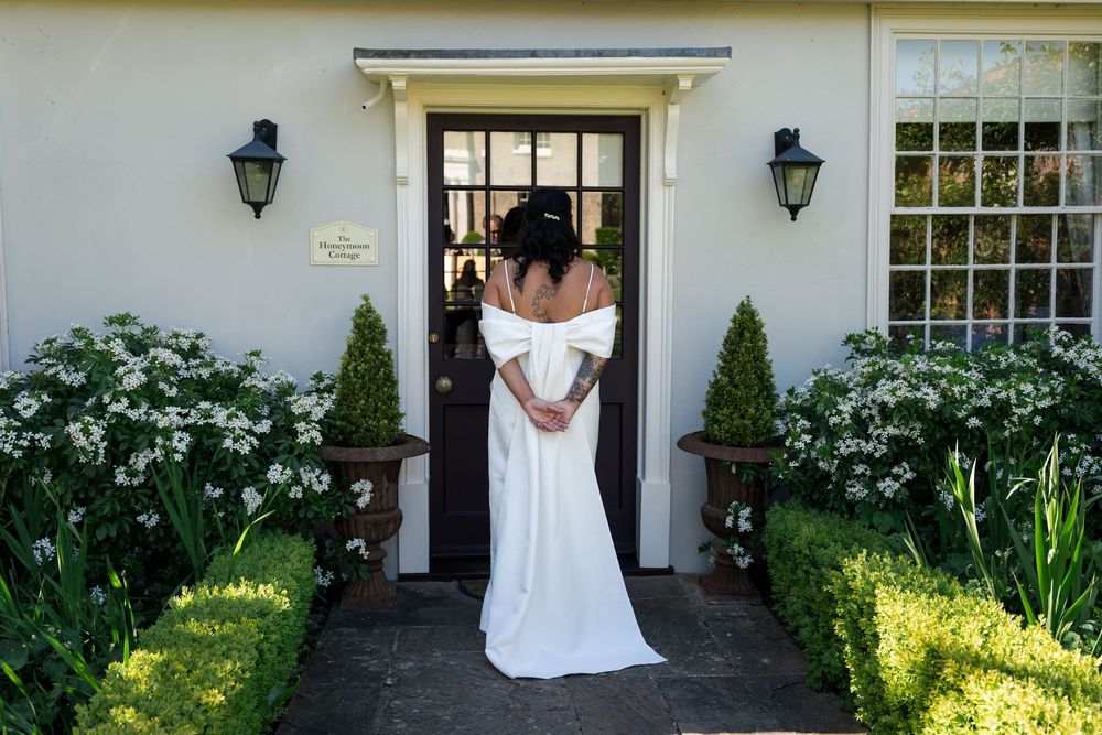 Bride in elegant white off-shoulder wedding gown standing at a charming cottage door surrounded by lush greenery and white flowers.