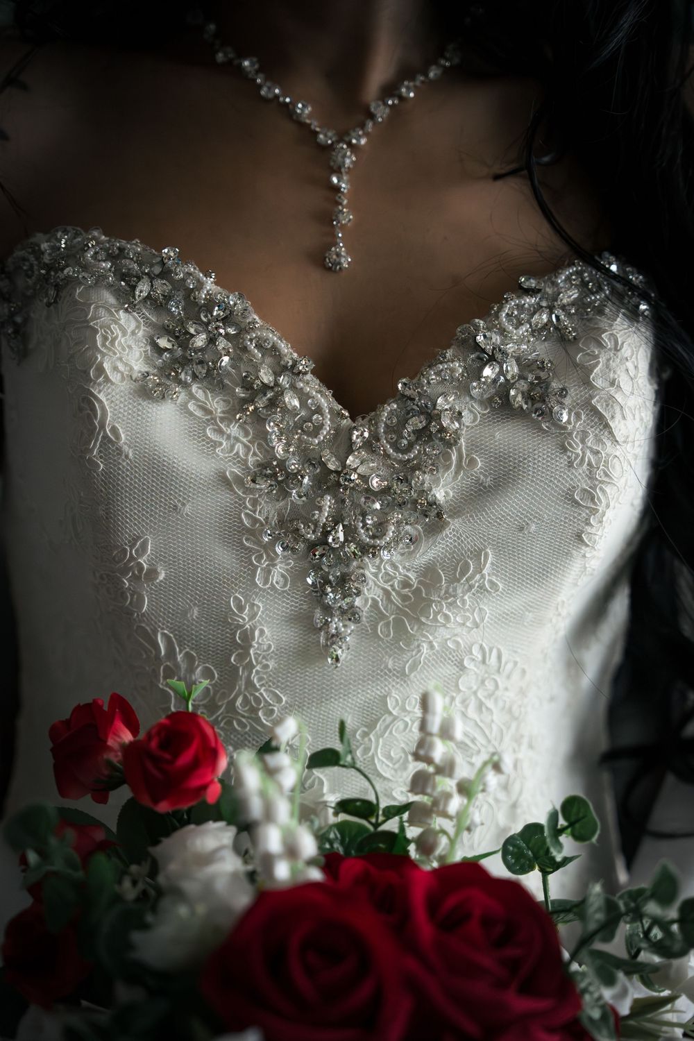 Bride in beaded sweetheart wedding gown with crystal necklace holding red rose bouquet.