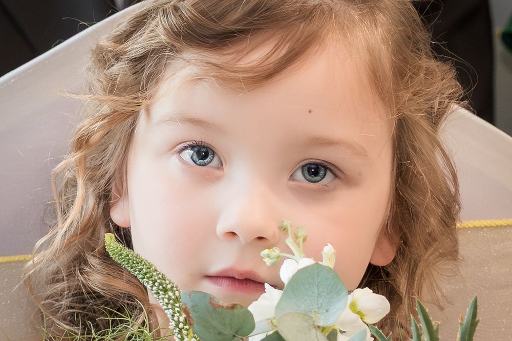 Close-up portrait of a young girl with blue eyes and curly hair holding white wedding flowers and eucalyptus greenery.