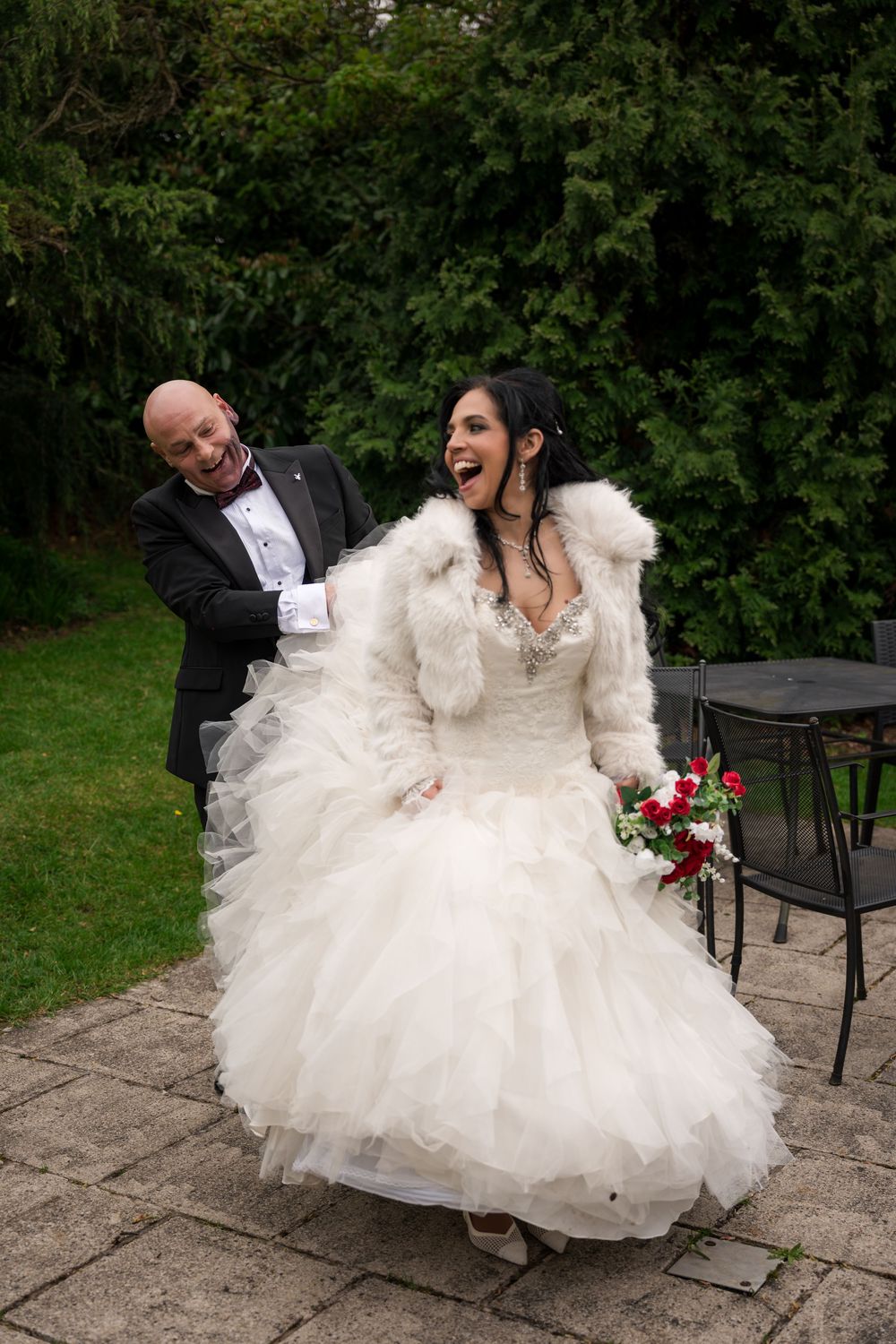 Joyful bride in white tulle gown and fur stole holds red bouquet while groom in tuxedo laughs behind her outdoors.