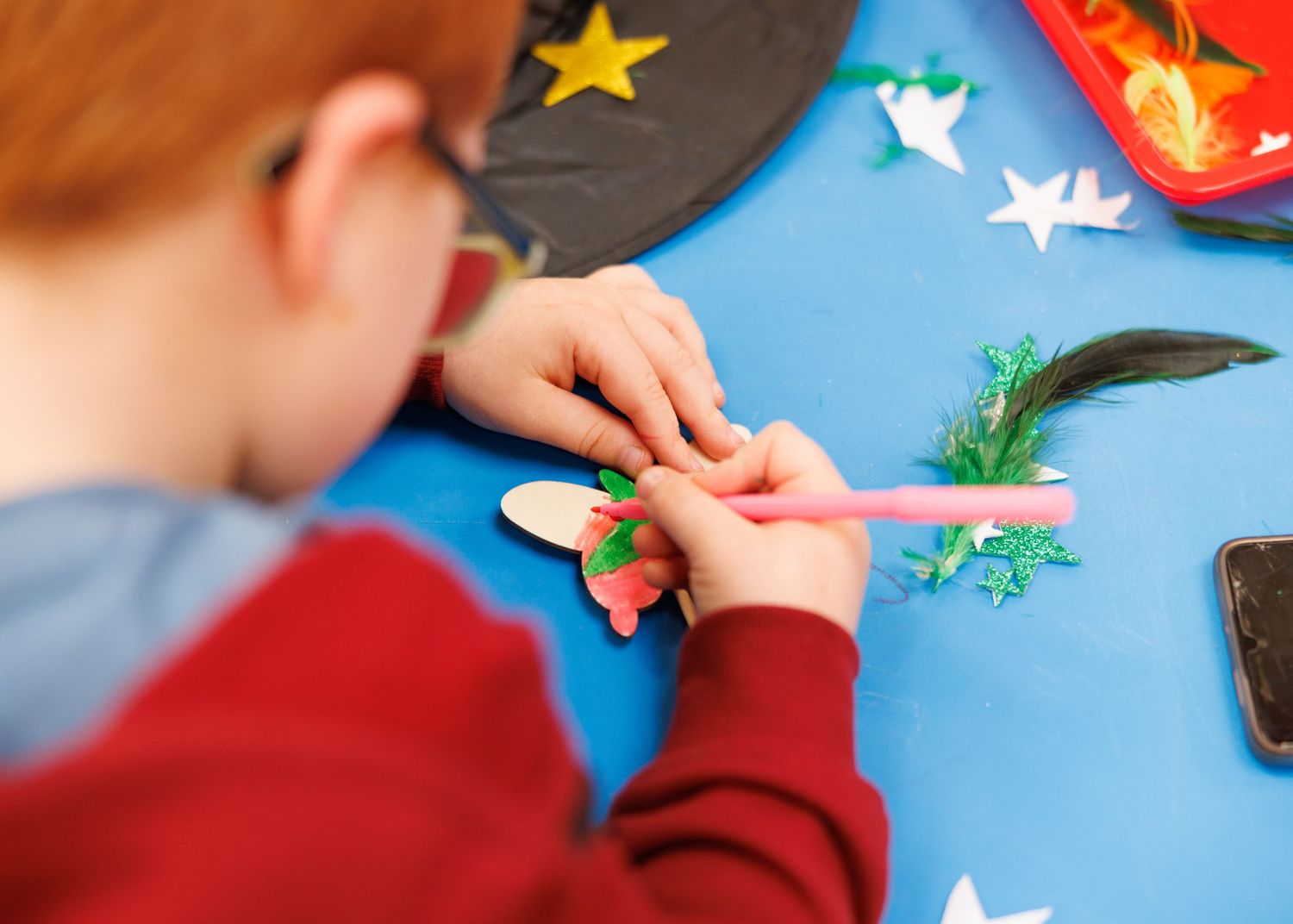 Small hands work on a craft project with colorful paper cutouts and decorative elements on a bright blue surface.