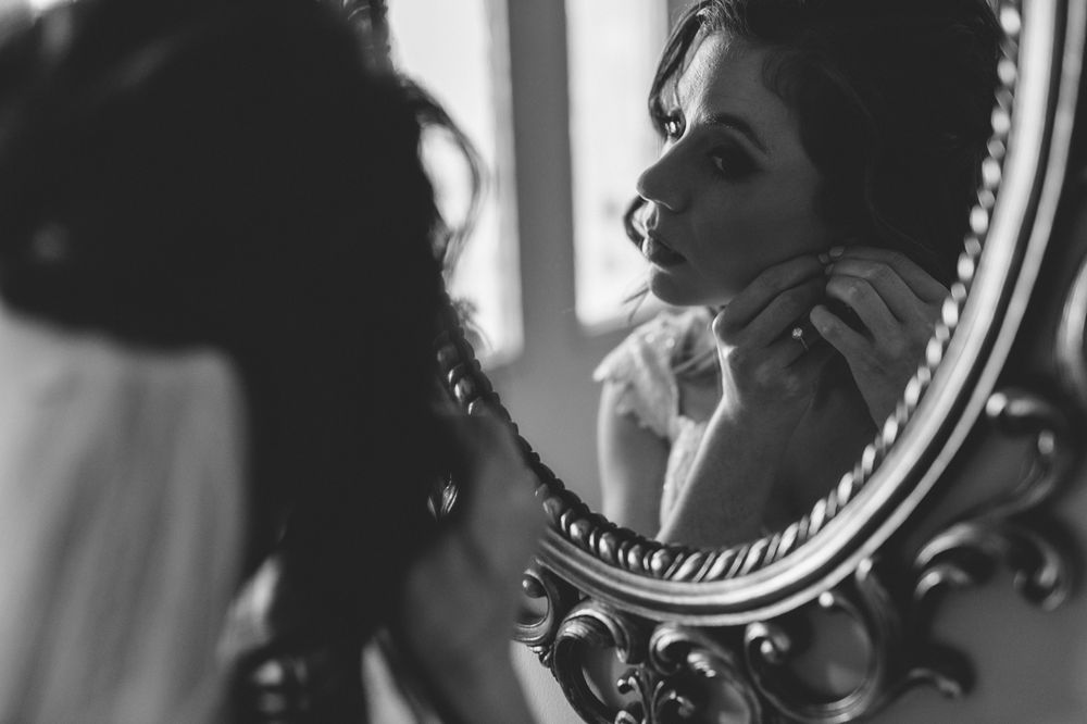 Black and white photo of a bride looking into a mirror putting her earrings on.