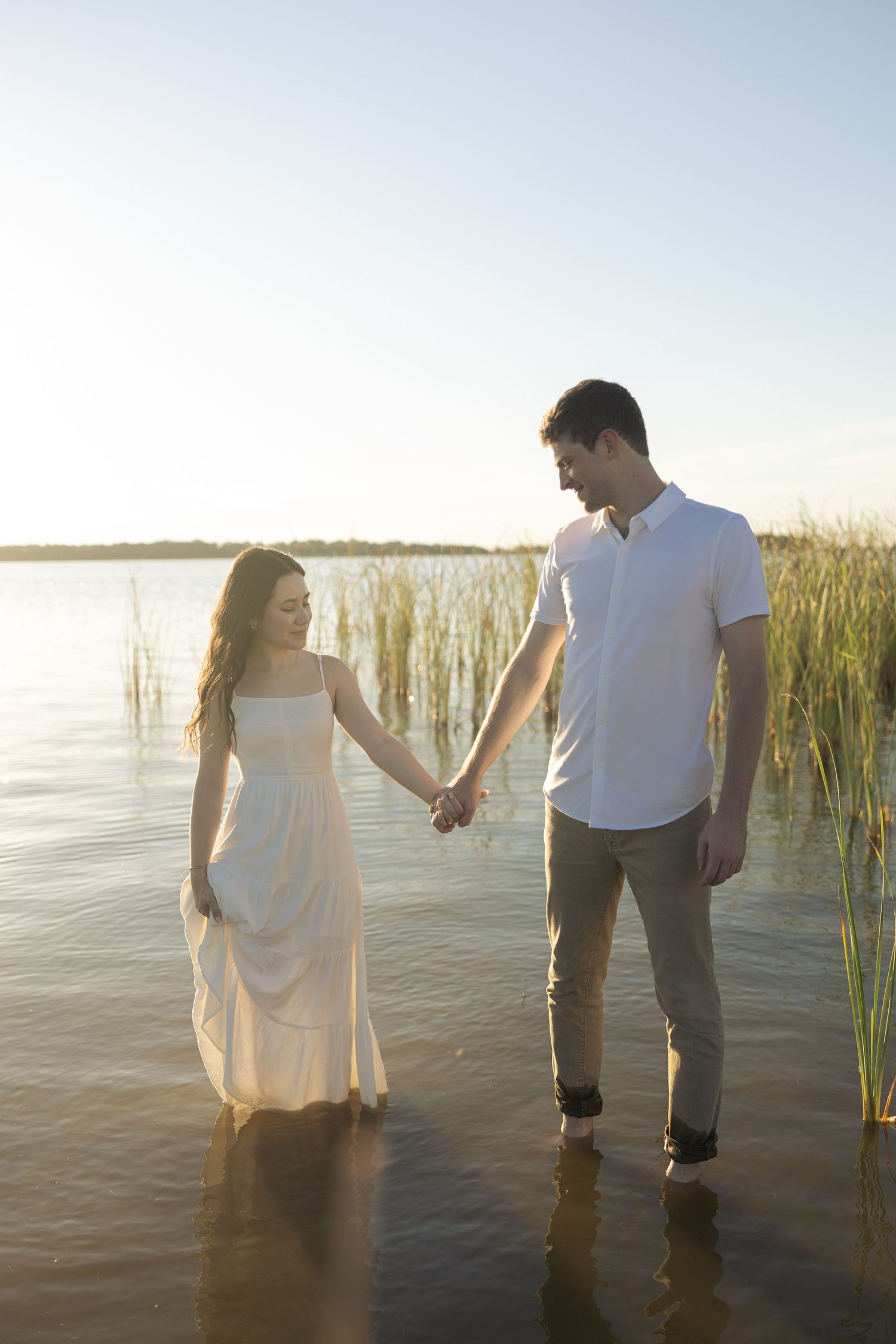 A couple stands in shallow water among reeds during a romantic outdoor photoshoot at sunset.