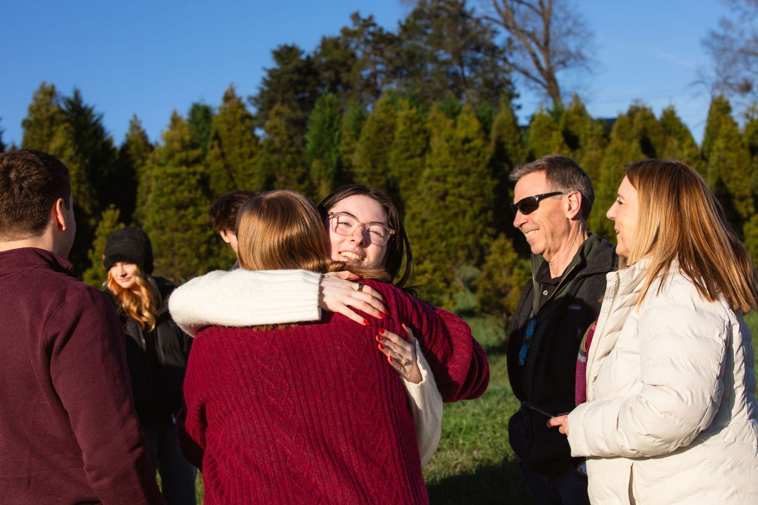 Family embracing and hugging at the tree farm after a surprise proposal.