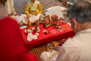 Traditional Indian wedding ceremony showing hands performing sacred rituals with flowers and offerings on red cloth.